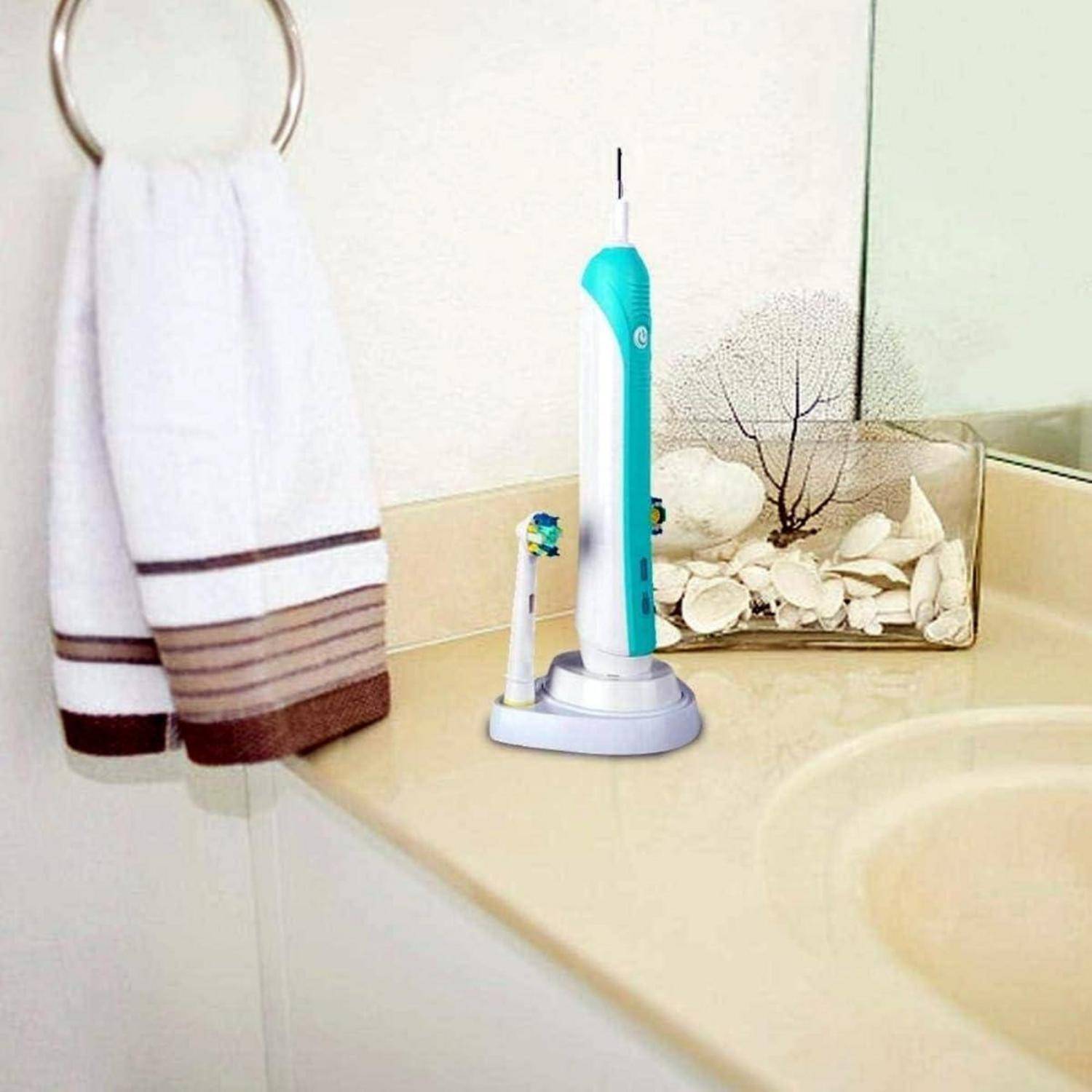 Elektrische Zahnbürste und Ersatzkopf auf einem Badezimmertresen neben einem Waschbecken, mit einem hängenden Handtuch und dekorativer Muscheldisplay.
