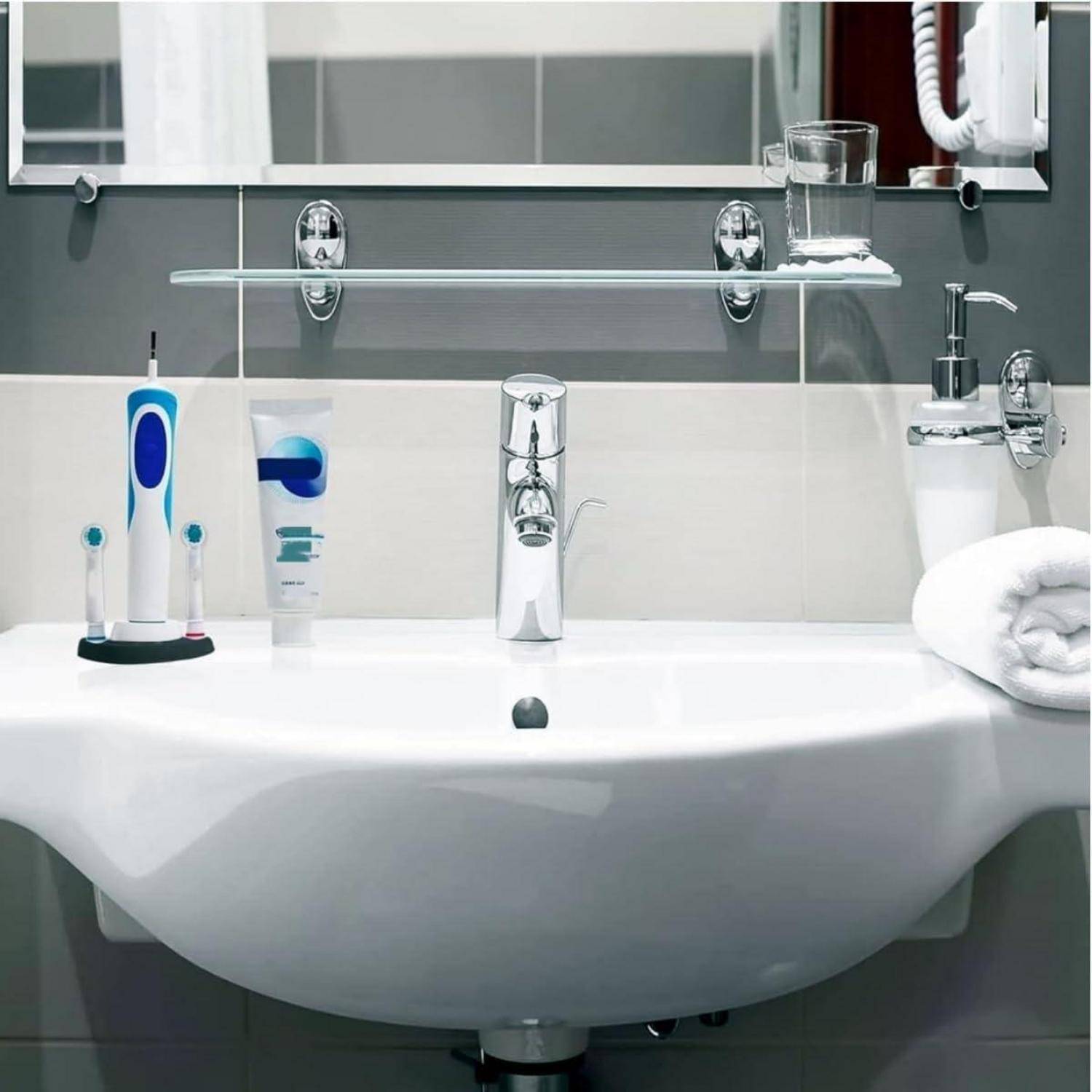 Modernes Badezimmerbecken mit Zahnbürste, elektrischer Zahnbürste, Zahnpasta, Seifenspender, Glas und aufgerolltem Handtuch auf der Arbeitsplatte und dem Regal.