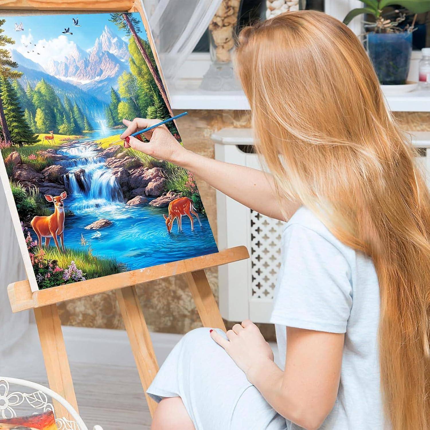 Eine Frau malt eine farbenfrohe Berglandschaft mit einem Wasserfall und Hirsche auf einer Staffelei.