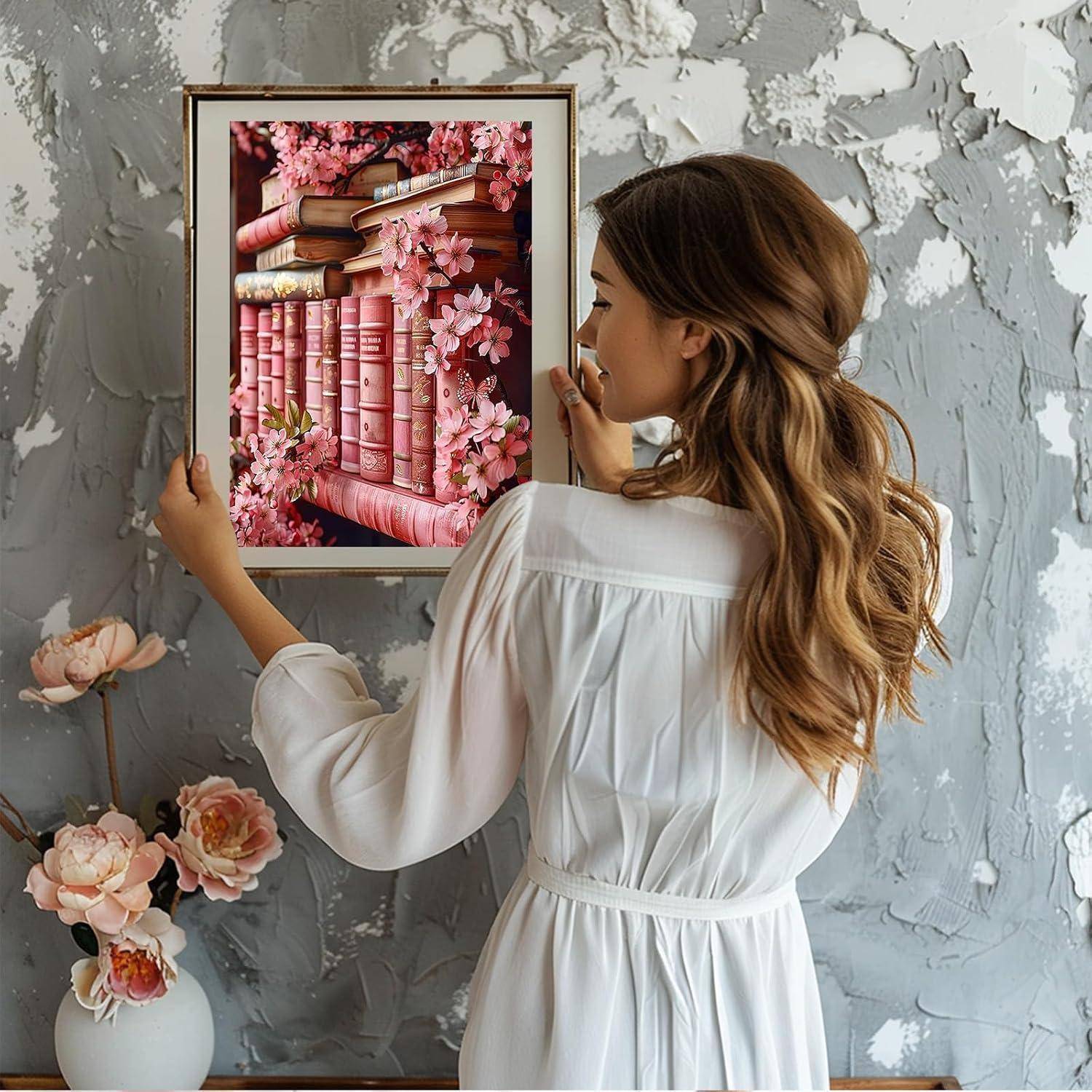 Eine Frau in einem weißen Kleid hängt ein gerahmtes rosa Kunstwerk mit Buch und Blüten an eine strukturierte graue Wand.