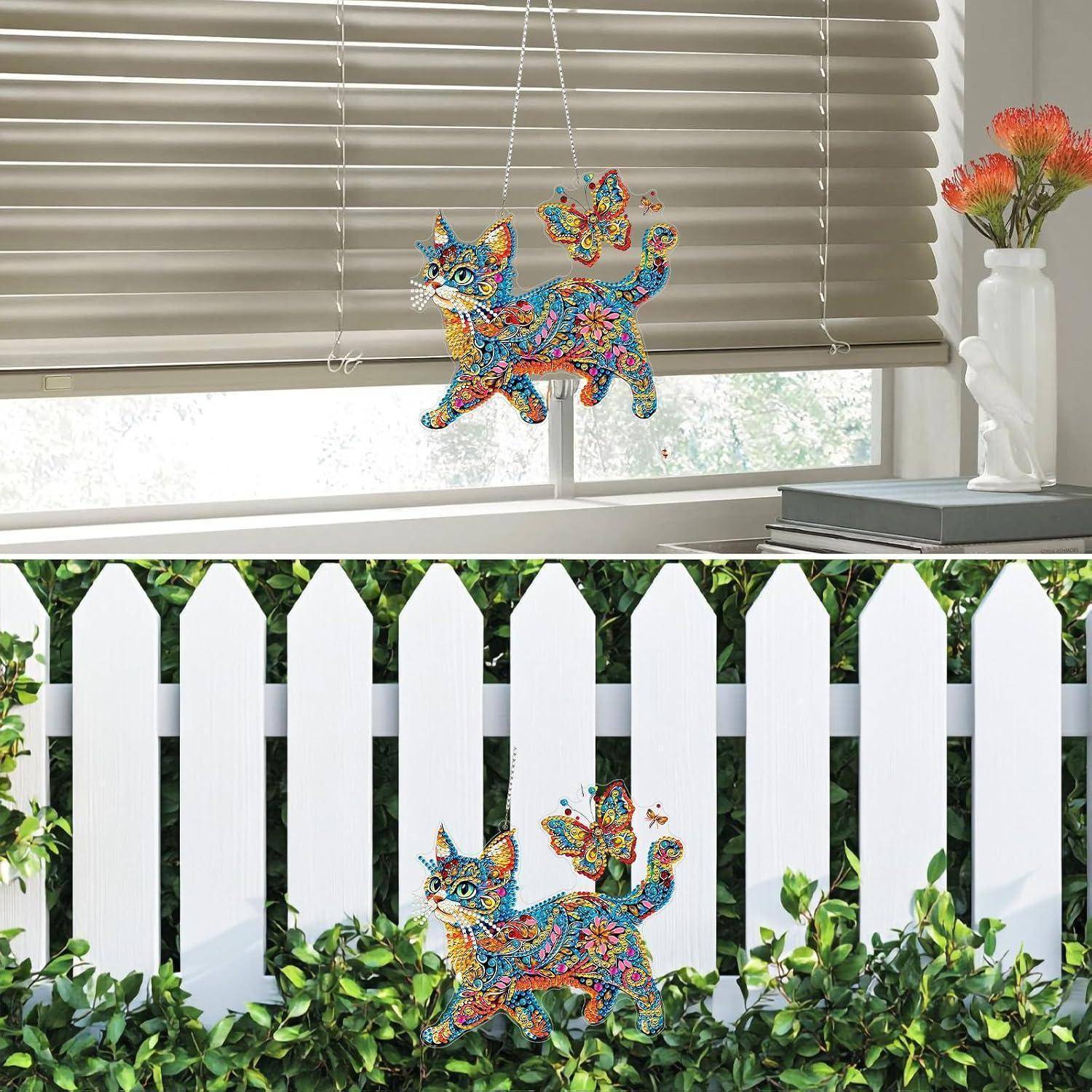 Bunter katzenhaft geformter Hängeschmuck, der in einem Fenster und auf einem weißen Lattenzaun angebracht ist, mit Schmetterlingen und Grünpflanzen in der Nähe.