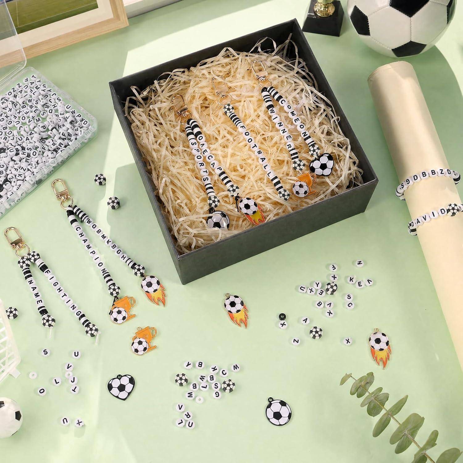 Fußball-themed personalisiertes Geschenkset mit einer schwarzen Schachtel Stifte und kleinen Charms, umgeben von Perlen, Schlüsselanhängern und Verpackung auf einer mintgrünen Oberfläche.