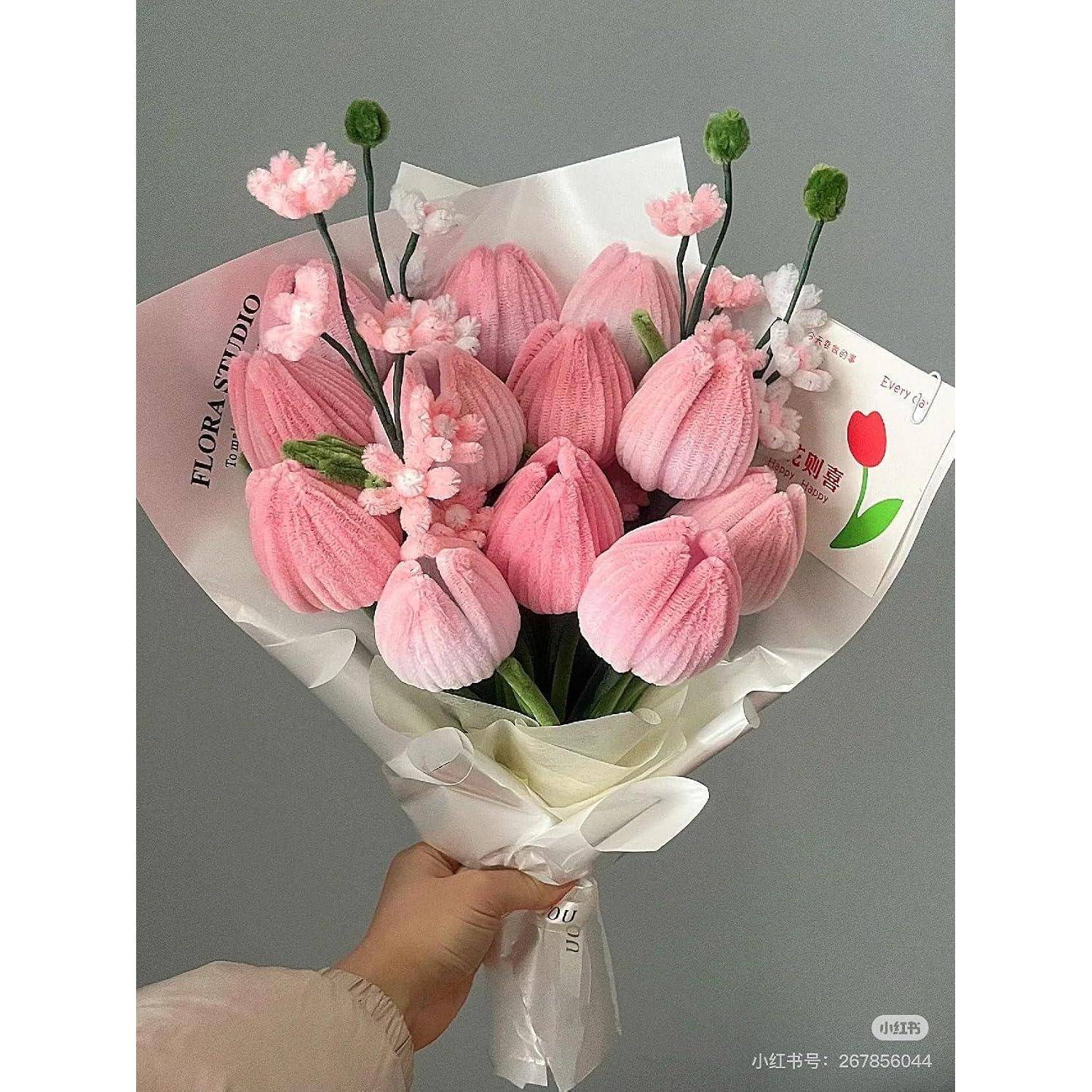 Eine Hand hält einen Strauß rosa tulpenähnlicher Blumen, die in weißes Papier eingewickelt sind, vor einem grauen Hintergrund.