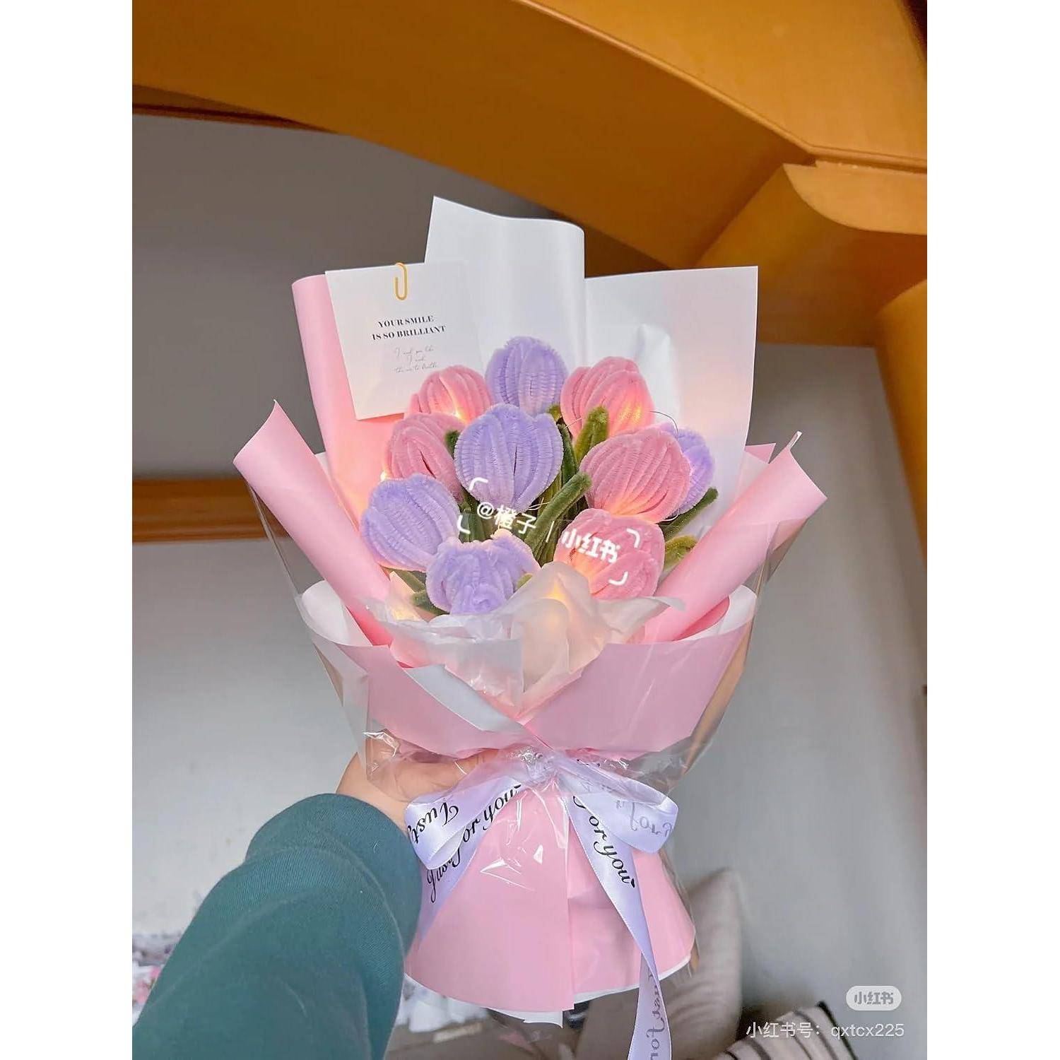Eine Hand hält einen zartem rosafarbenen Blumenstrauß, der in rosa- und weißem Papier eingewickelt ist, mit Lavendel- und rosafarbenen blütenförmigen Dekorationen und einer kleinen Notizk­arte, die daran befestigt ist.