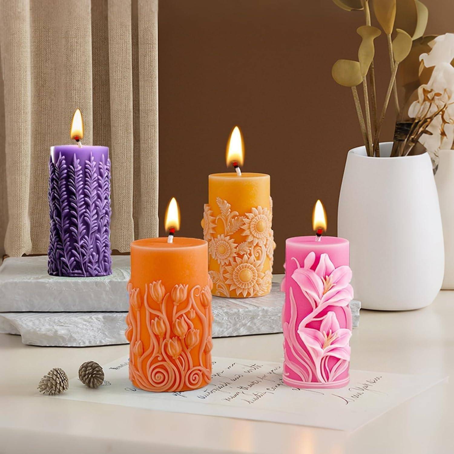 Vier dekorative beleuchtete Säulenkerzen in Lila, Orange, Gelb und Rosa mit geschnitzten Blumenmotiven auf einer Tischplatte neben einer weißen Vase und Blumen.