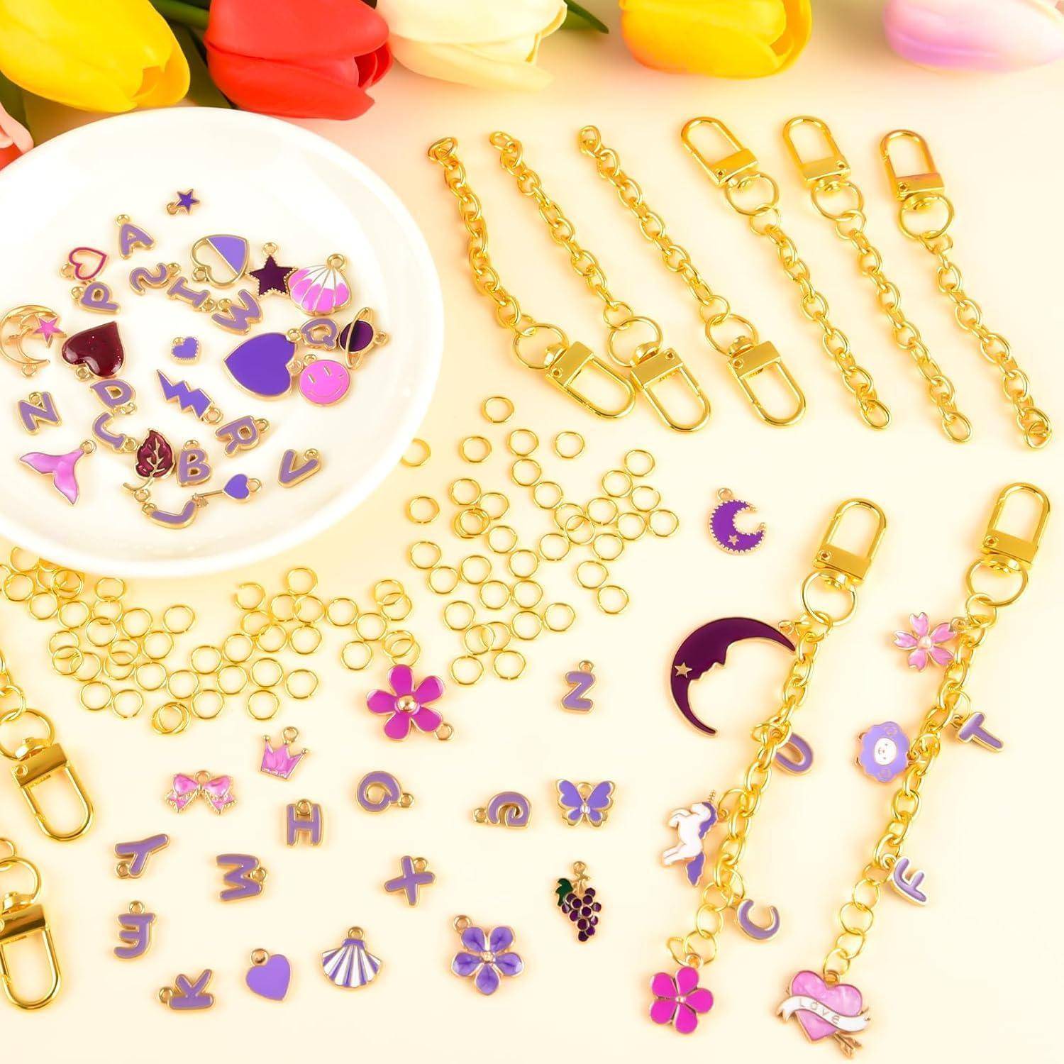 Sortierte Goldjuwelierarbeit-Zubehörteile und violette Emaille-Anhänger, einschließlich Ketten, Verschlüsse, Sprungringe und dekorative Buchstaben, auf einer hellen Oberfläche angeordnet.