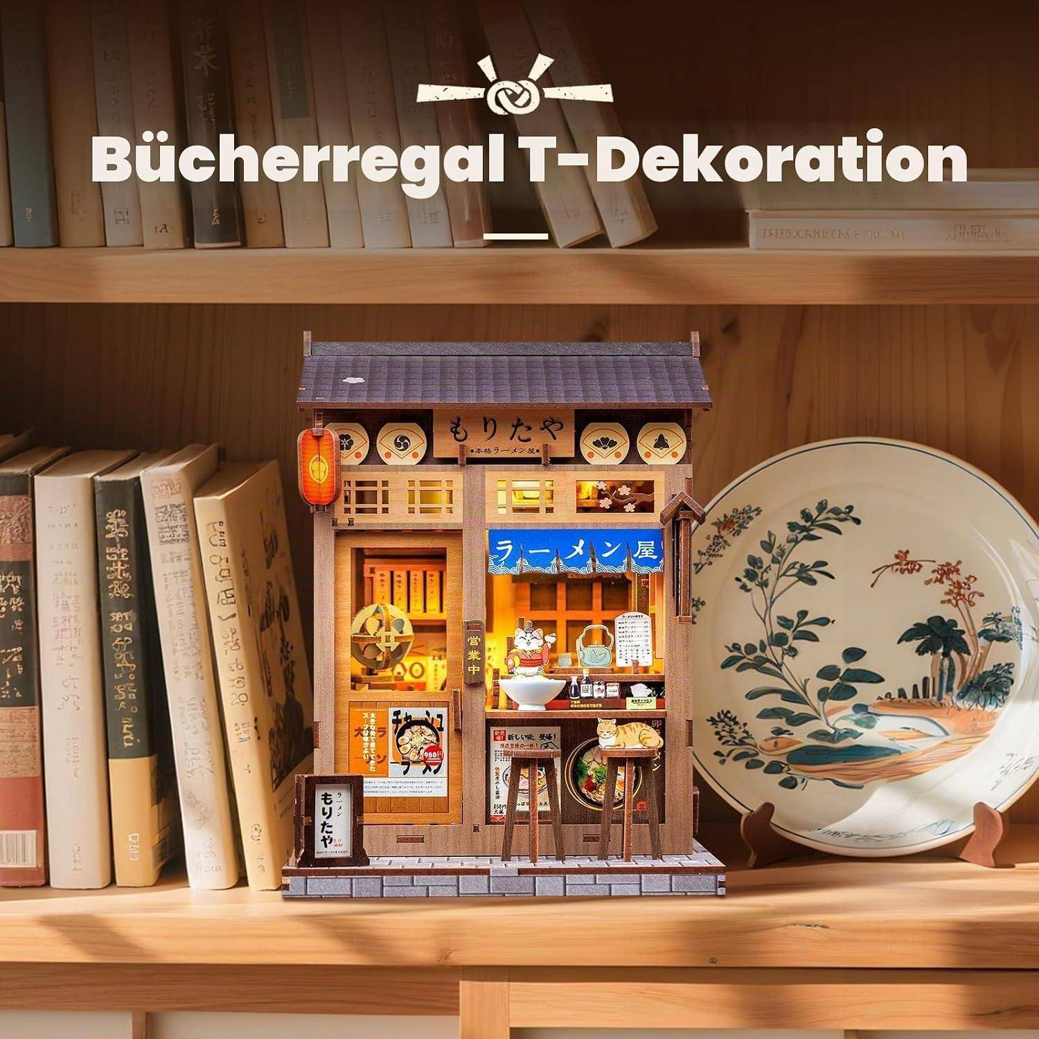 Ein miniaturelles japanisches Ramen-Shop-Modell, das auf einem Holzbücherregal ausgestellt ist, flankiert von Büchern und einem dekorativen Blütenteller, mit dem deutschen Text „Bücherregal T-Dekoration