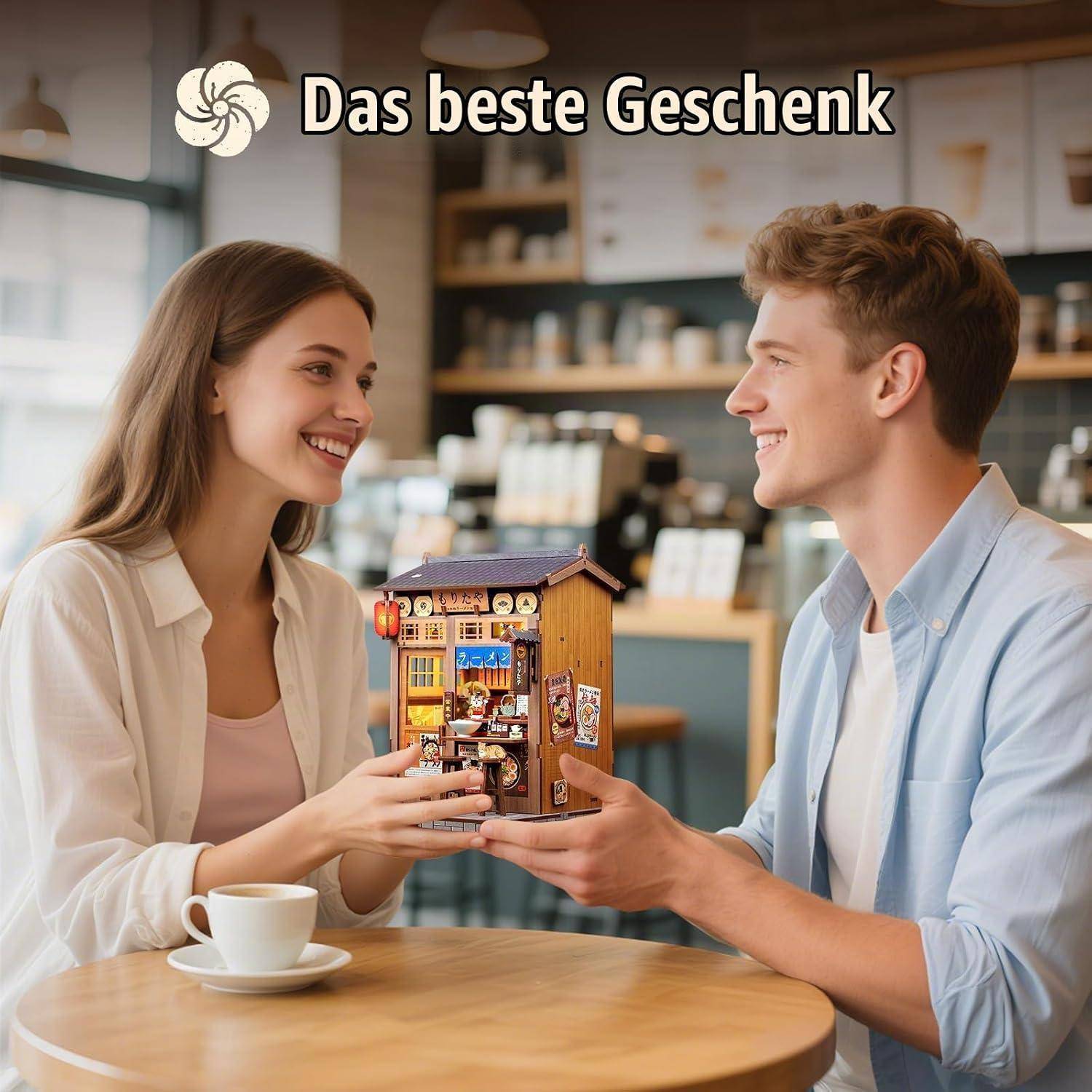 Junges lachendes Paar in einem Café tauscht eine kleine dekorative Geschenkbox aus; Text liest „Das beste Geschenk