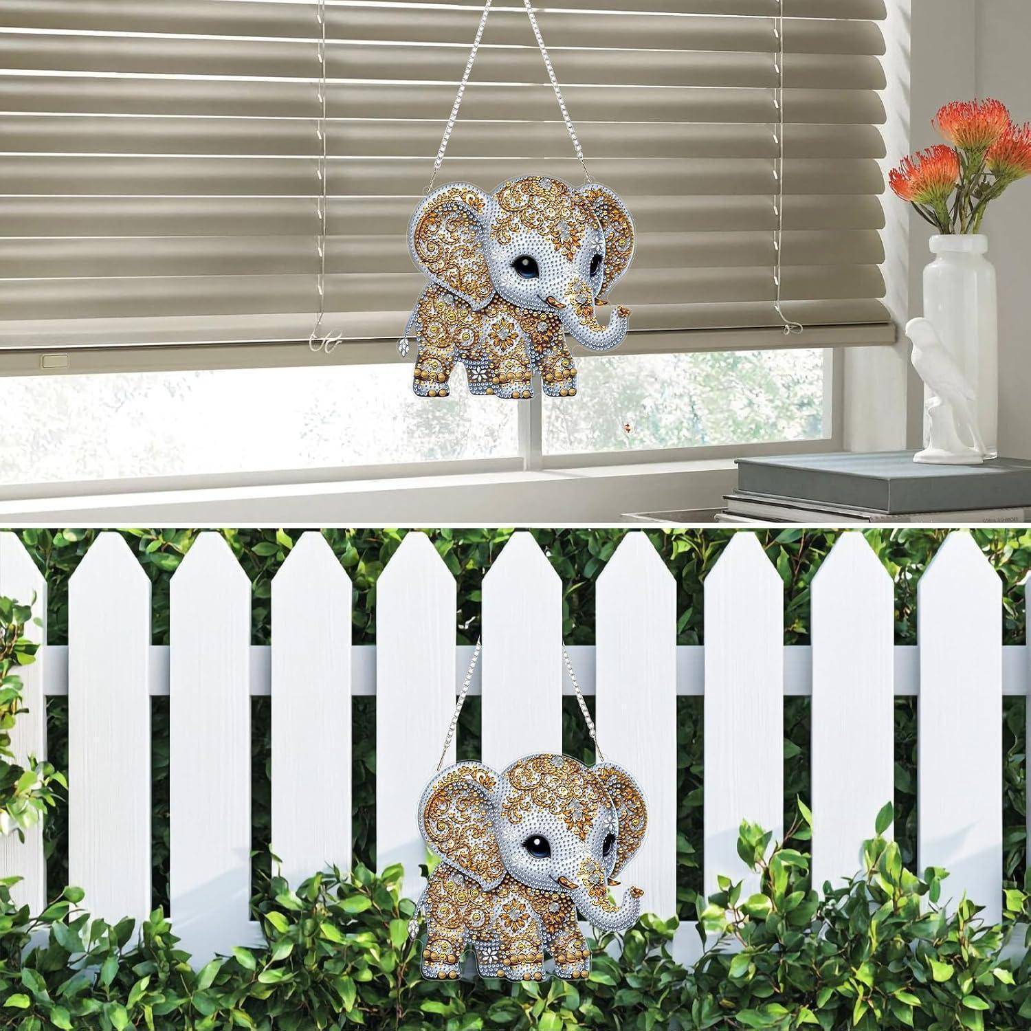 Dekorativer Elefantenschmuck mit Juwelen, der an einem Fenster hängt; wird erneut auf einem weißen Lattenzaun im Freien zwischen Grünpflanzen gezeigt.