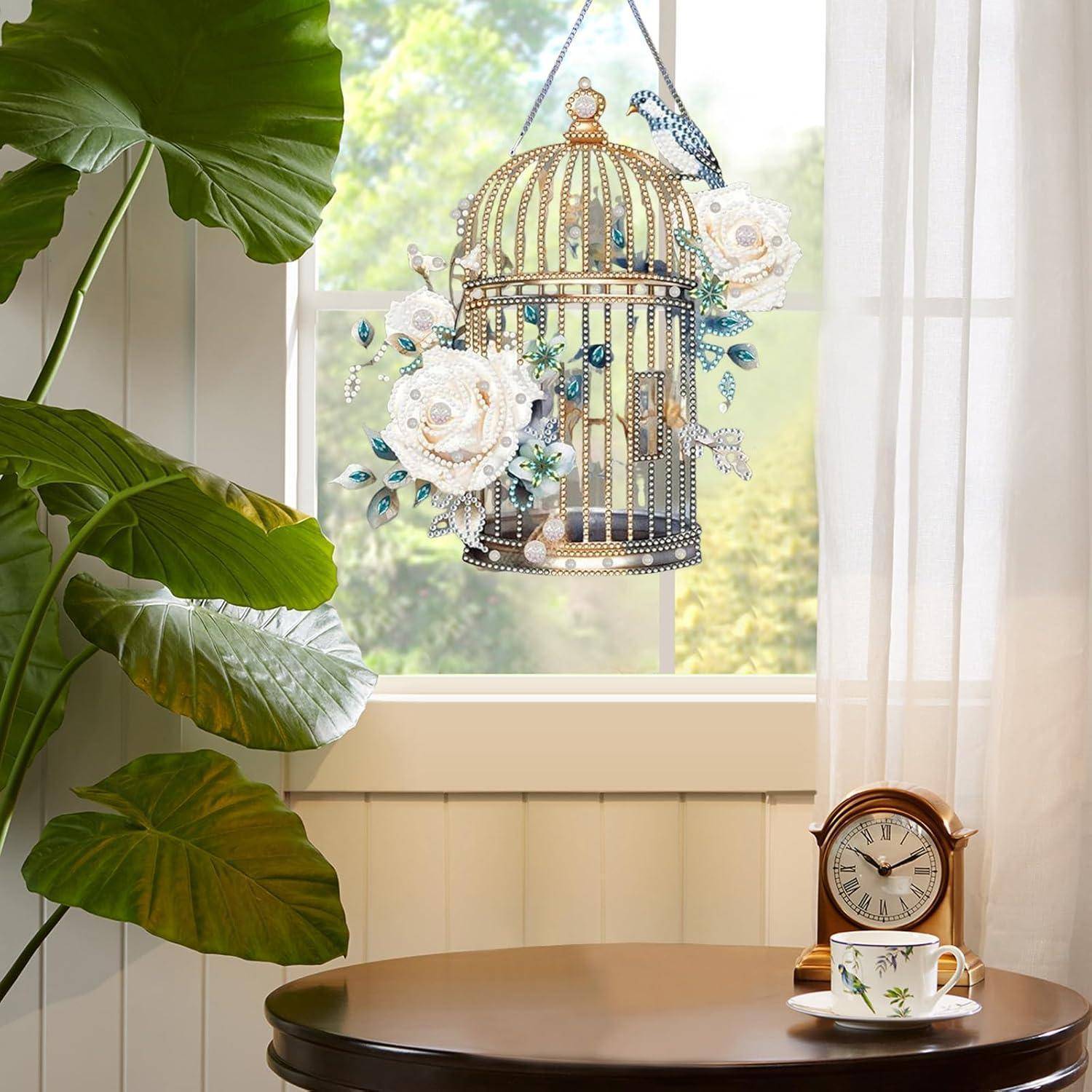 Dekoratives Vogelkäfig-Ornament mit weißen Blüten hängt in einem hellen Fenster neben einer Topfpflanze, mit einer Teetasse und einer Uhr auf einem kleinen Tisch.