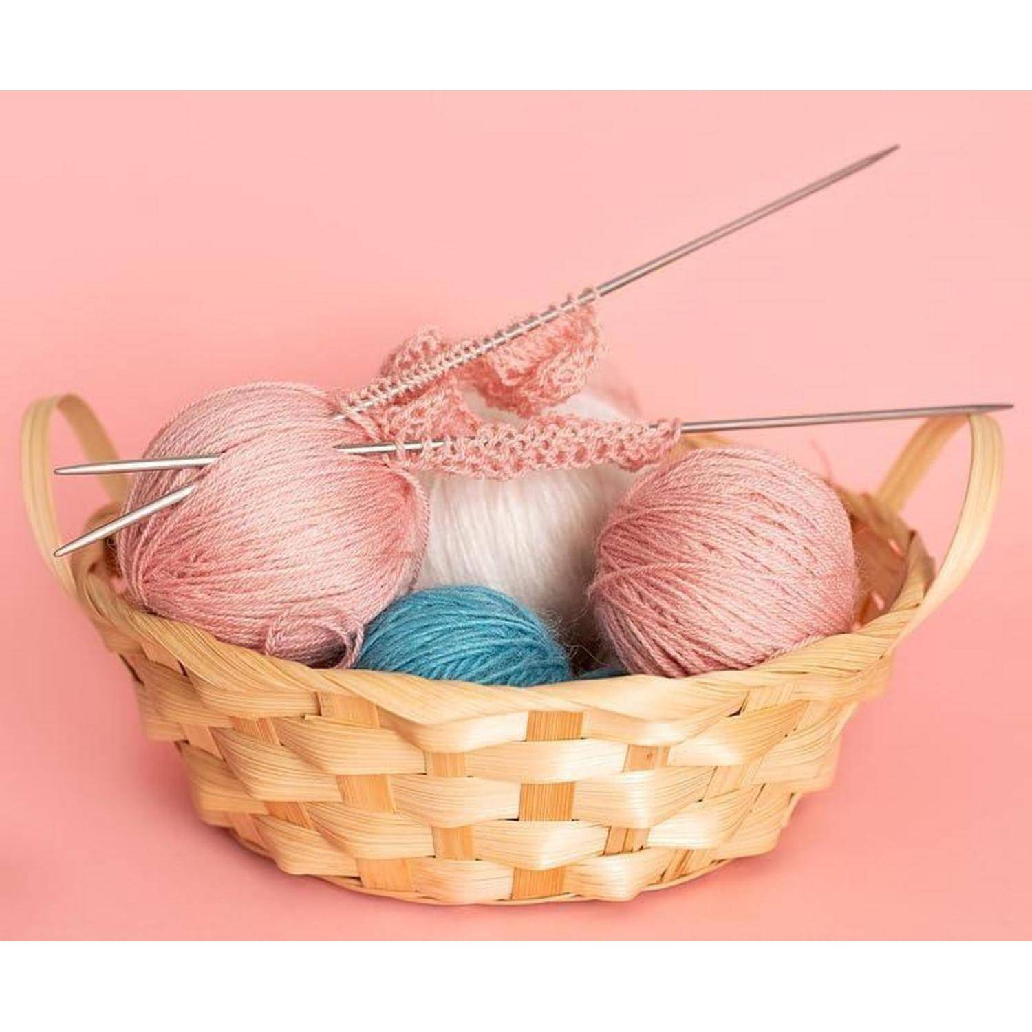 Ein Weidenkorb enthält pastellrosa, weiße und blaue Wollknäuel mit Stricknadeln und einem kleinen rosa gestrickten Stück auf rosa Hintergrund.