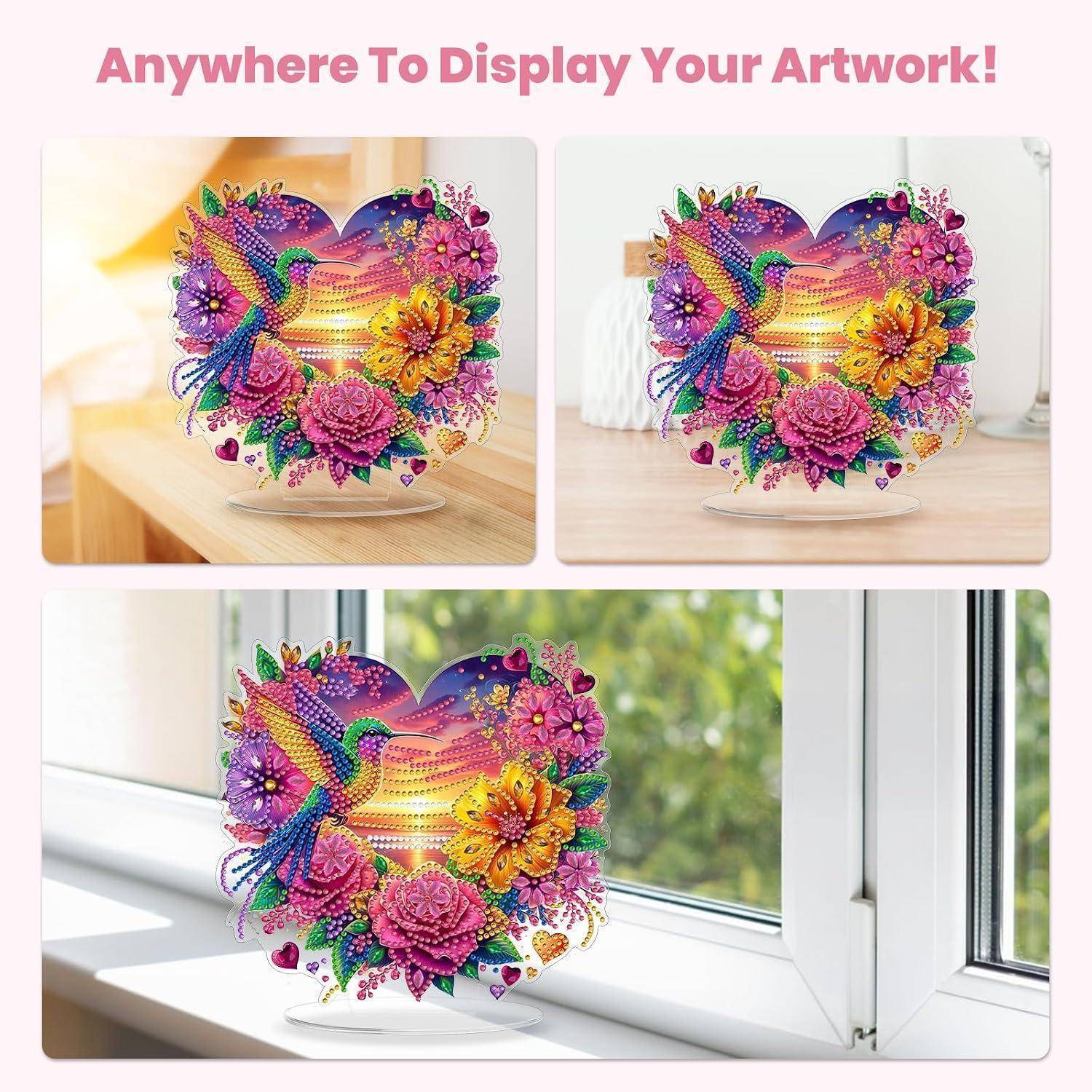 Ein herzförmiges, farbenfrohes Blumen- und Kolibri-Kunstwerk, das in drei Umgebungen präsentiert wird: auf einem Tisch, einem Regal und einer Fensterbank, mit der Beschriftung „Überall, um dein Kunstwerk auszustellen!"