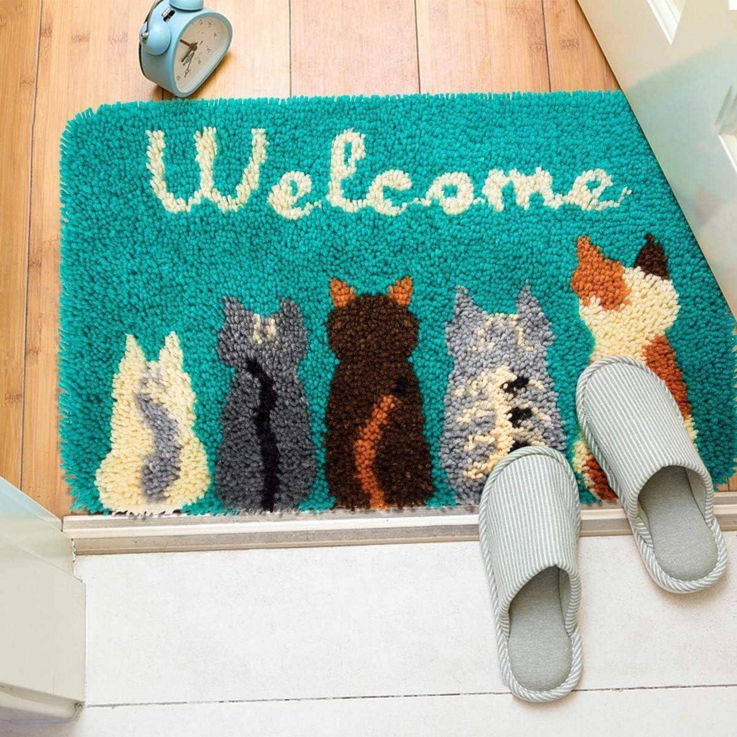 Türkisfarbene 'Willkommen'-Fußmatte mit fünf Katzenabbildungen neben einem Paar grauer Hausschuhe in einer Türöffnung.