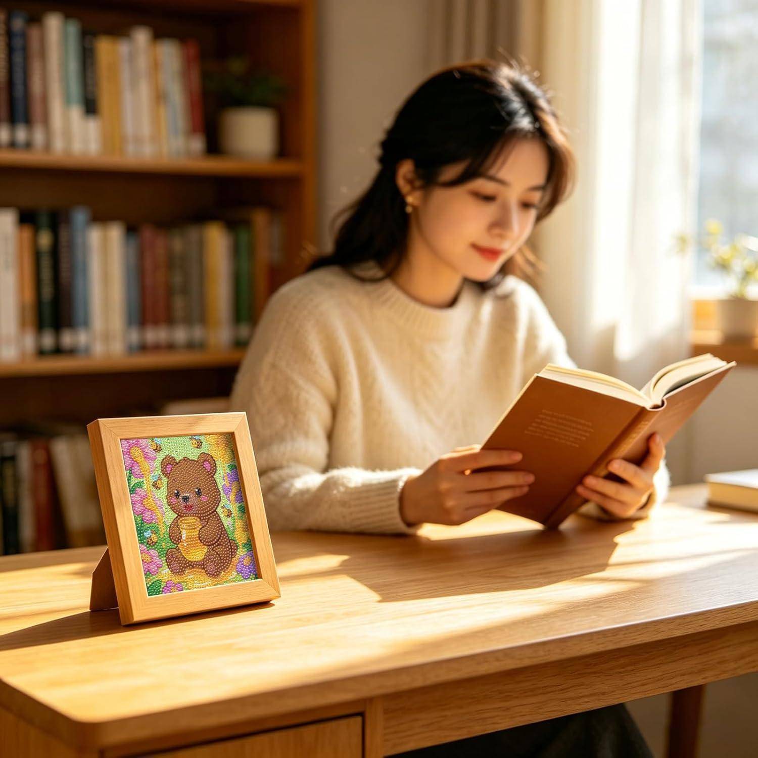 Eine Frau liest ein Buch an einem Holztisch in einem sonnendurchfluteten Zimmer, mit einem gerahmten Teddybärenbild im Vordergrund und Bücherregalen hinter ihr.