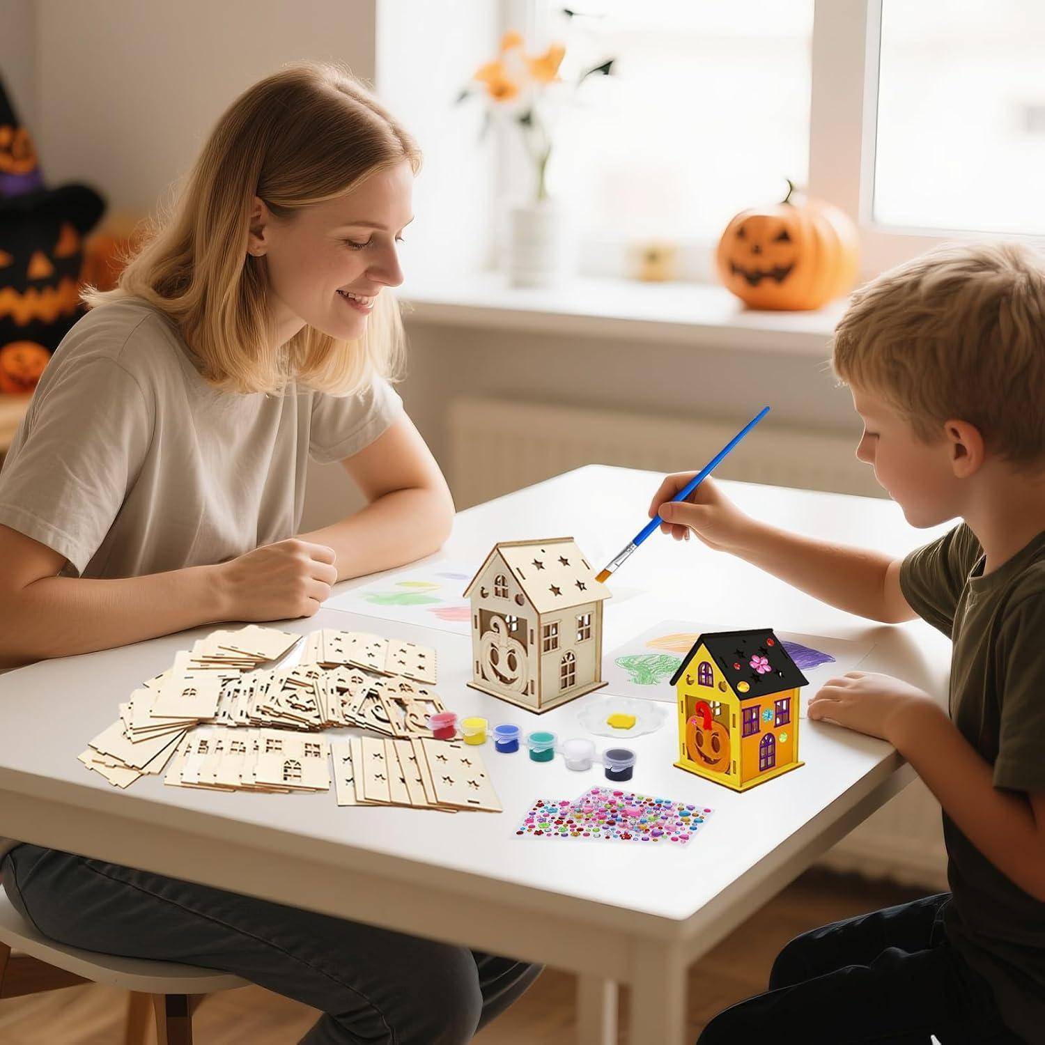 Frau und kleiner Junge bemalen kleine hölzerne Hausmodelle auf einem Tisch mit Kunstmaterialien in einem hellen Raum mit Halloween-Dekorationen.