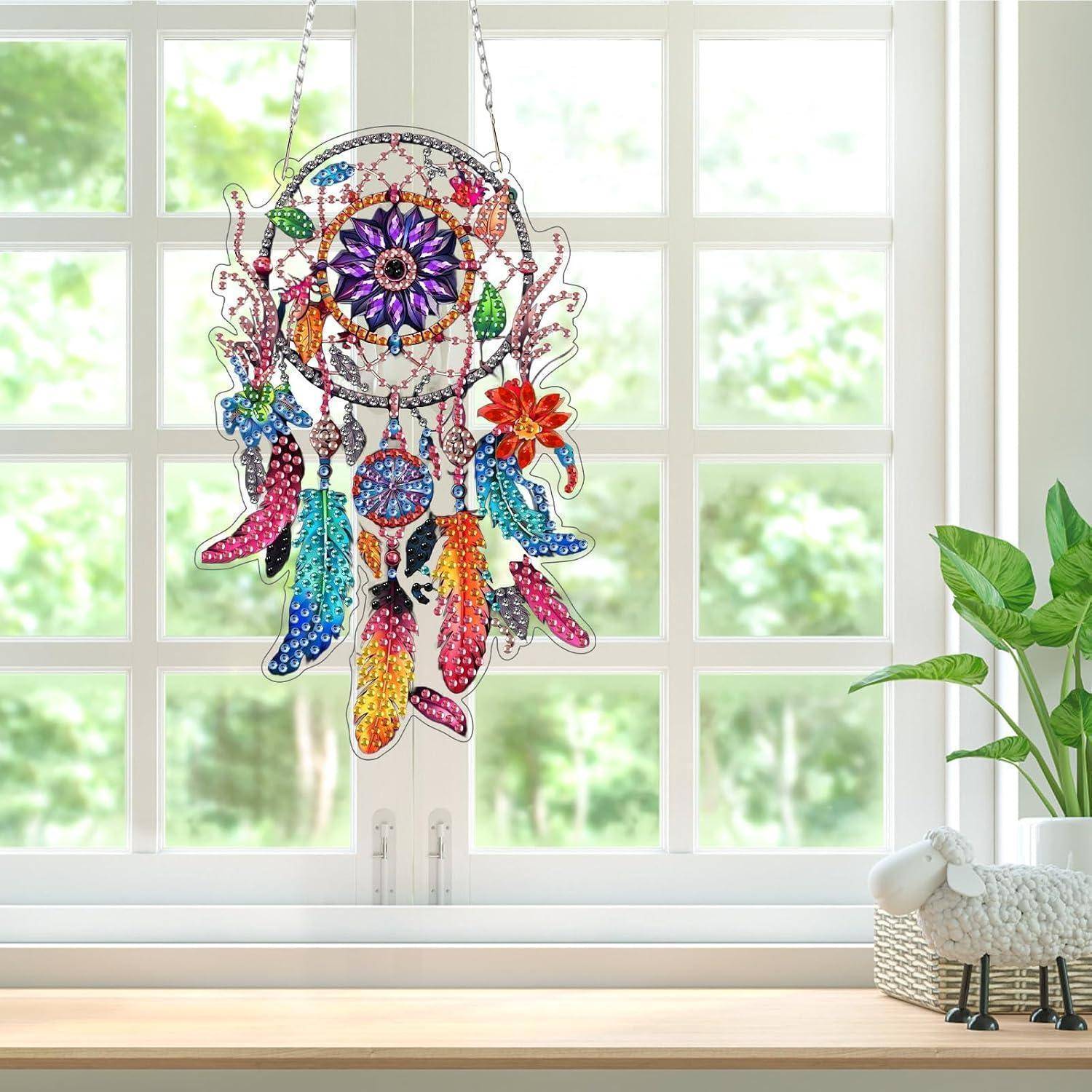 Farbenfroher traumfänger-ähnlicher Hängeschmuck mit Federn und Blumendetails, der in einem hellen Fenster neben einer Topfpflanze ausgestellt ist.