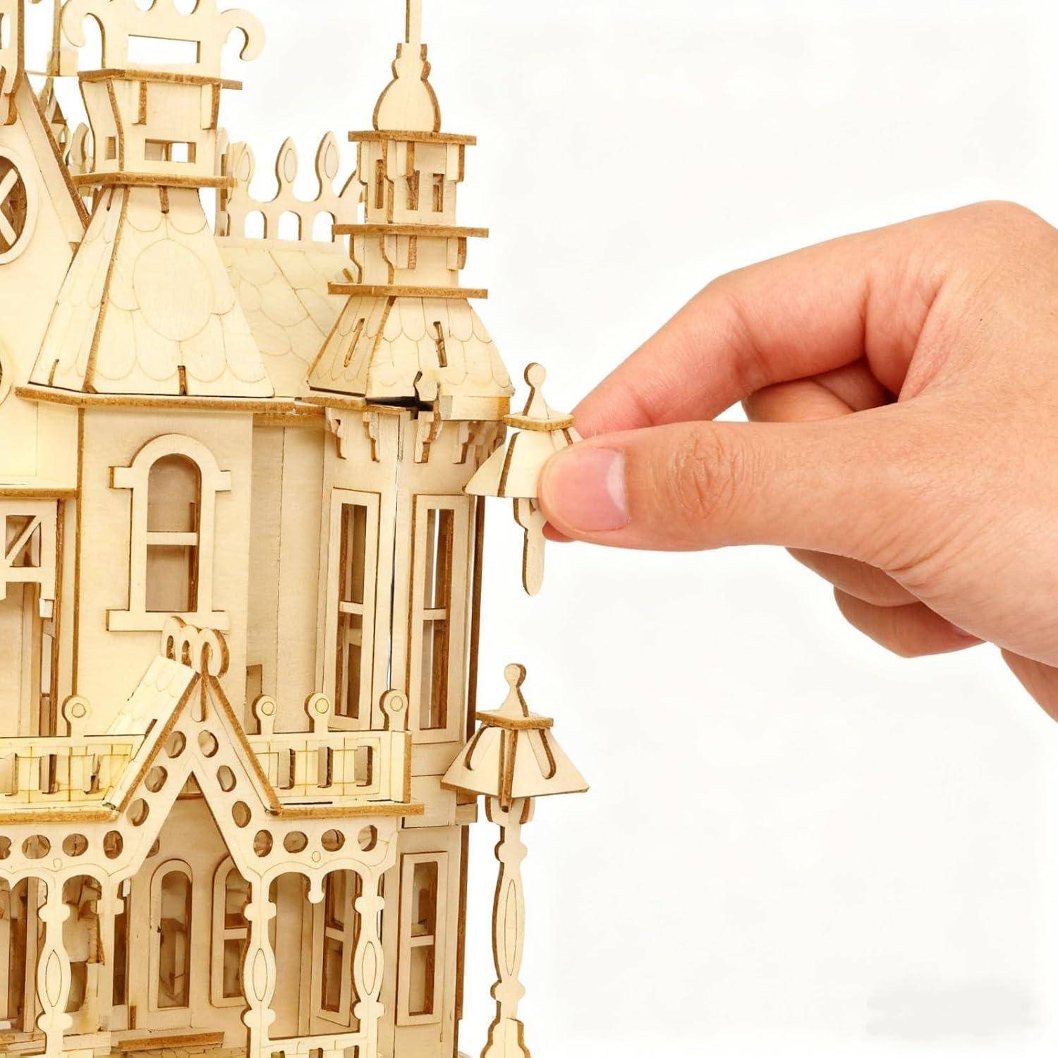 Eine Hand, die ein kleines hölzernes Ornament auf ein miniaturarchitektonisches Modell setzt, mit einer detaillierten Hausfassade auf der linken Seite.