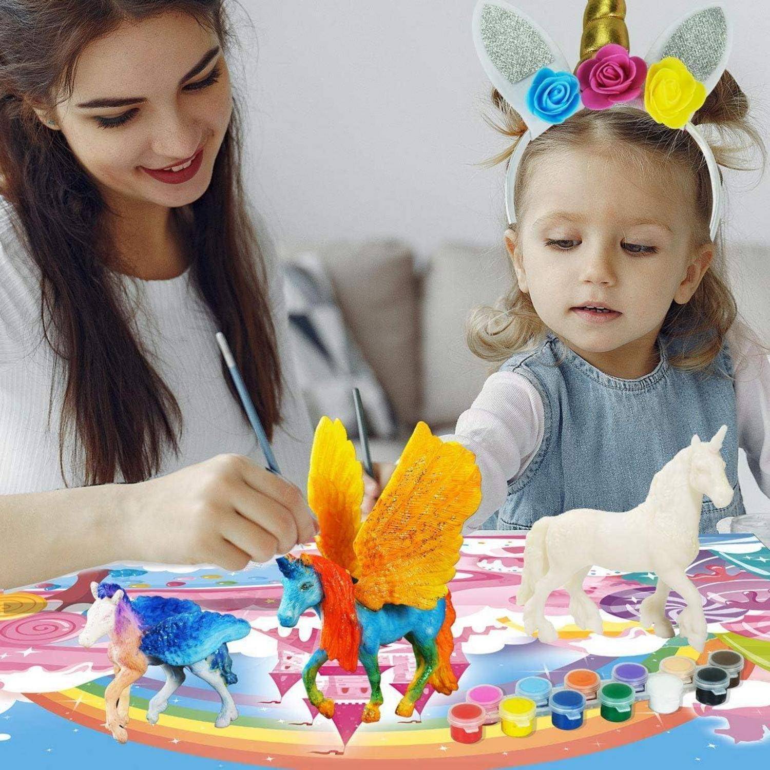 Eine Frau hilft einem jungen Mädchen, bunte Spielzeug-Einhörner und ein geflügeltes Pferd an einem Tisch während einer Bastelaktivität anzumalen.