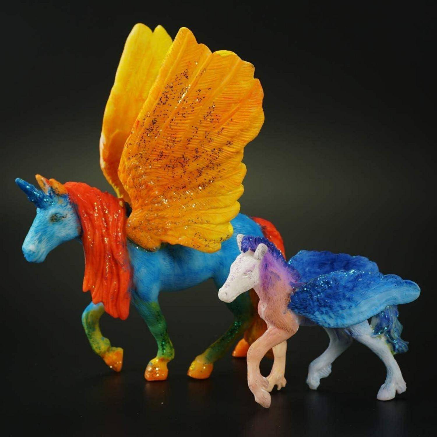 Zwei bunte geflügelte Einhorn-Figuren, eine größere in Blau mit orangefarbenen Flügeln und eine kleinere in Weiß-und-Blau, auf schwarzem Hintergrund angeordnet.