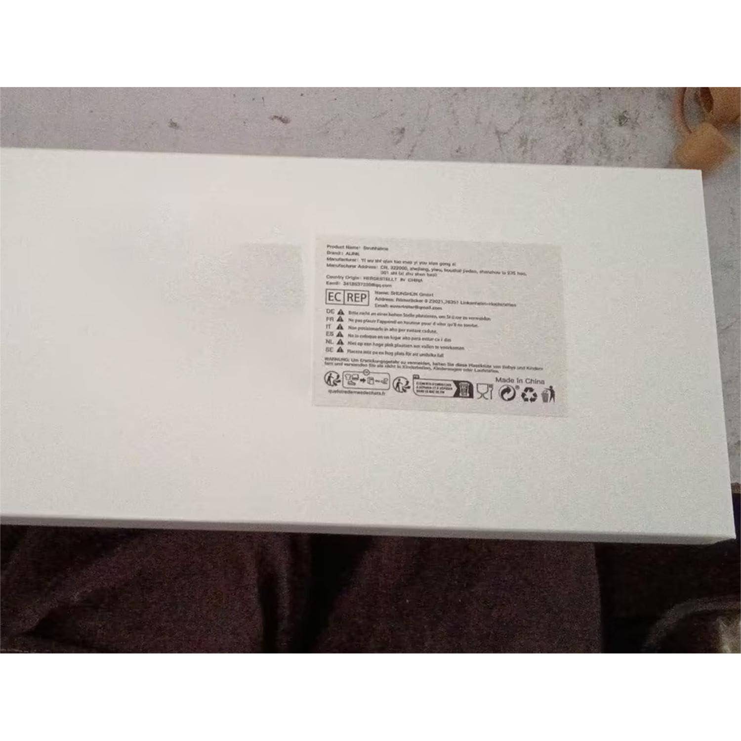 Weiße Produktbox mit einem kleinen aufgedruckten Etikett und Sicherheitssymbolen oben, fotografiert in Innenräumen auf dem Schoß einer Person.