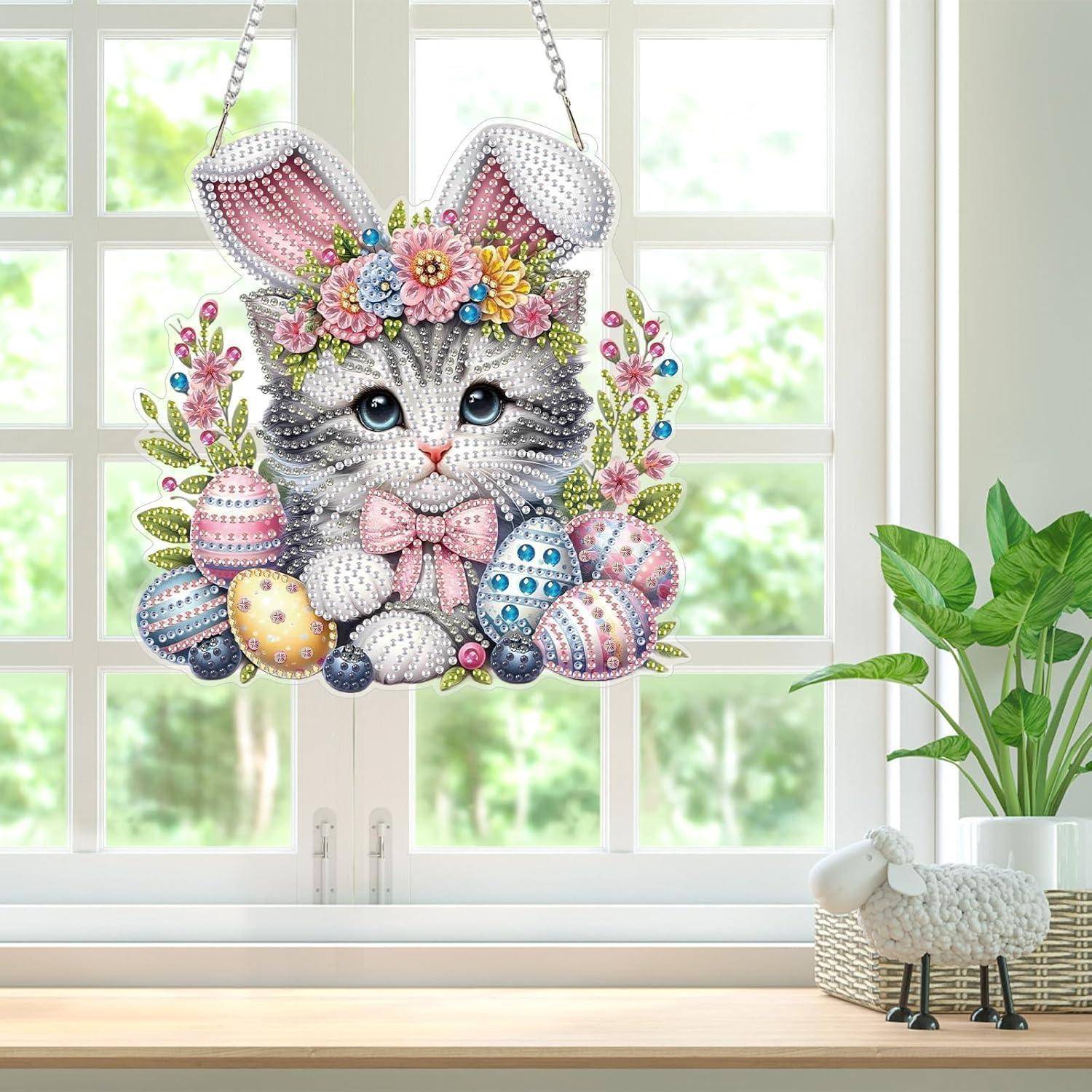 Ein funkelndes Diamant-Kunst-Hasen-Osterdeko mit Blumen und bemalten Eiern hängt in einem hellen Fenster über einer Fensterbank mit einer Pflanze und einer kleinen Schaf-Figur.