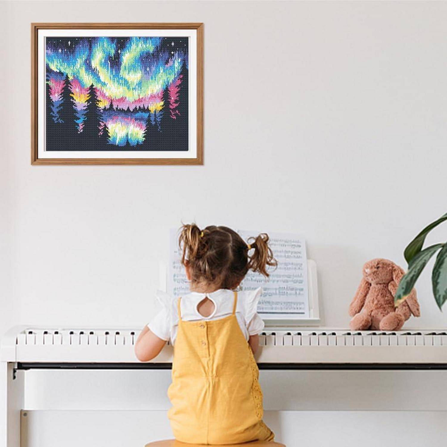 Kleines Kind, das an einem weißen Klavier sitzt, blickt auf Notenblätter in einem hellen Zimmer mit einer gerahmten farbenfrohen Landschaftsaufnahme darüber und einem Teddybär auf dem Klavier.