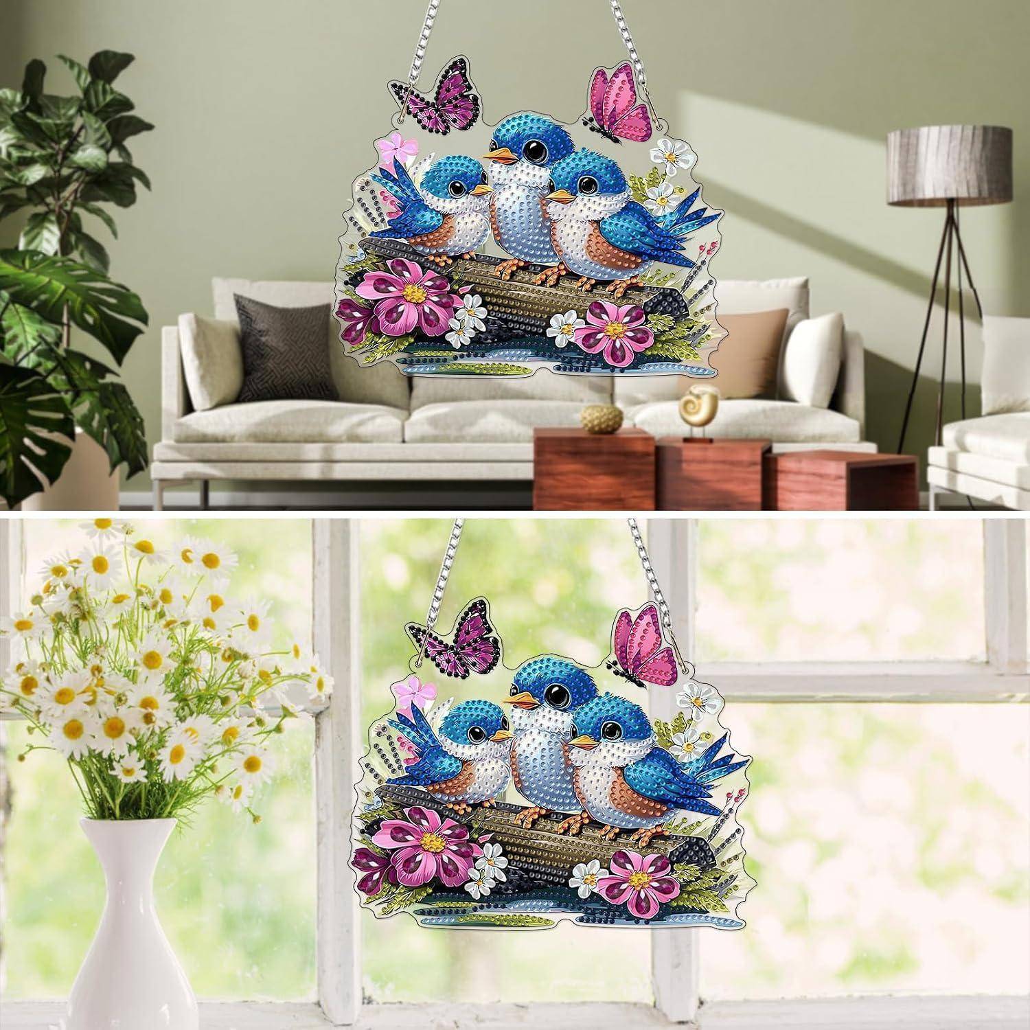 Farbenfrohe hängende Vogelverzierung mit drei blauen Vögeln, Schmetterlingen und Blumen, gezeigt in Innenräumen und in einem hellen Fenster.