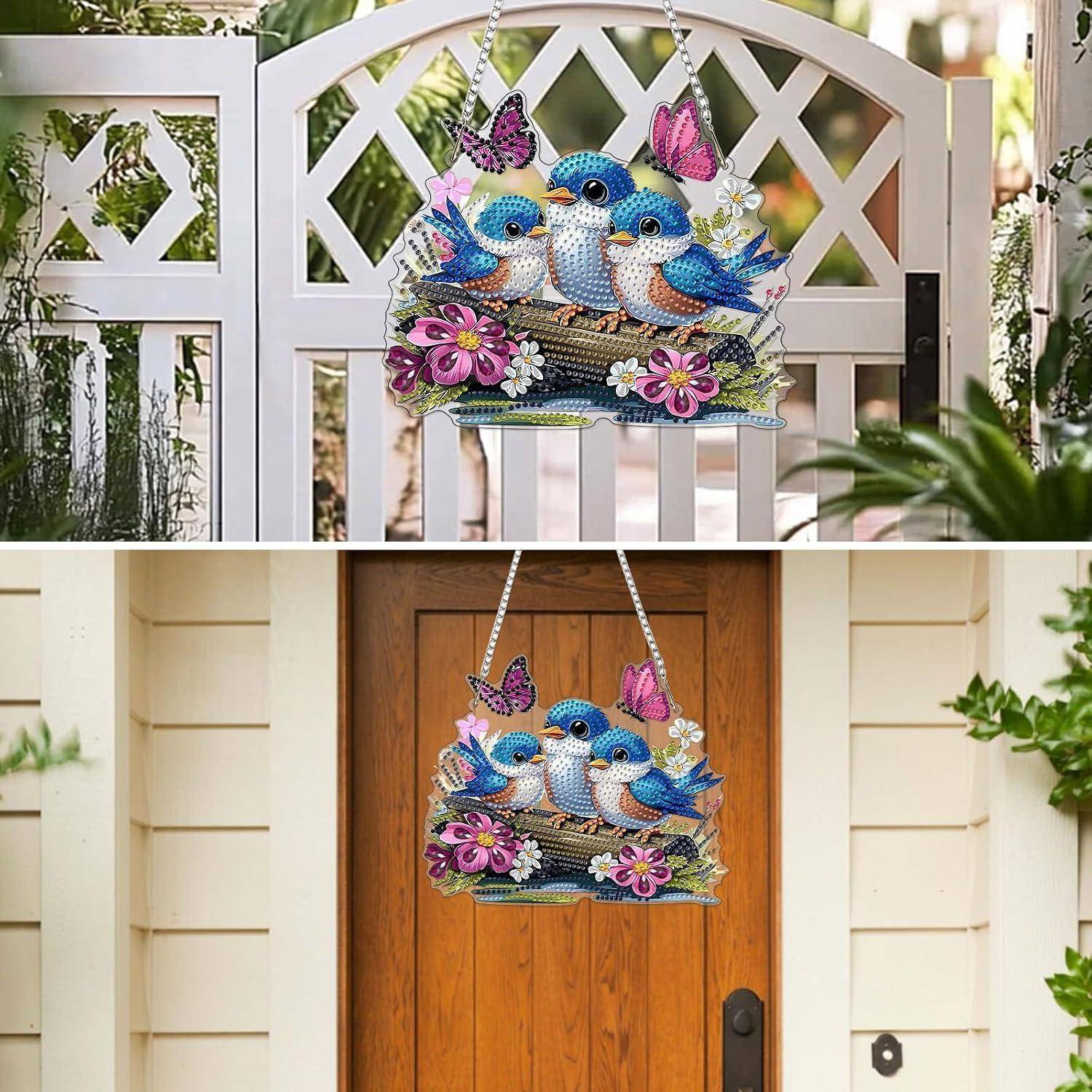 Buntes Vogel- und Schmetterlingsgartenzeichen, das an einem weißen Tor und einer Haustür hängt und drei blaue Vögel mit rosa Blumen zeigt.