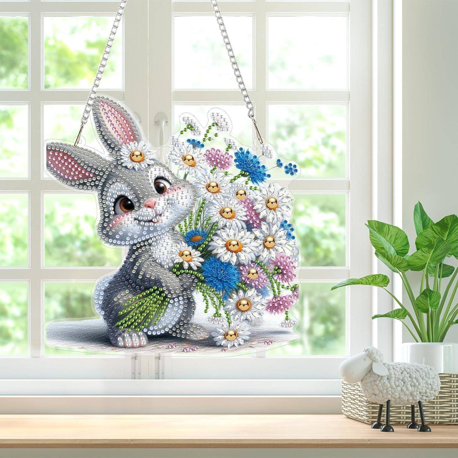 Diamantmalerei eines lächelnden Kaninchens mit Blumen, das in einem hellen Fenster hängt, mit einer kleinen dekorativen Schaffigur auf der Fensterbank.