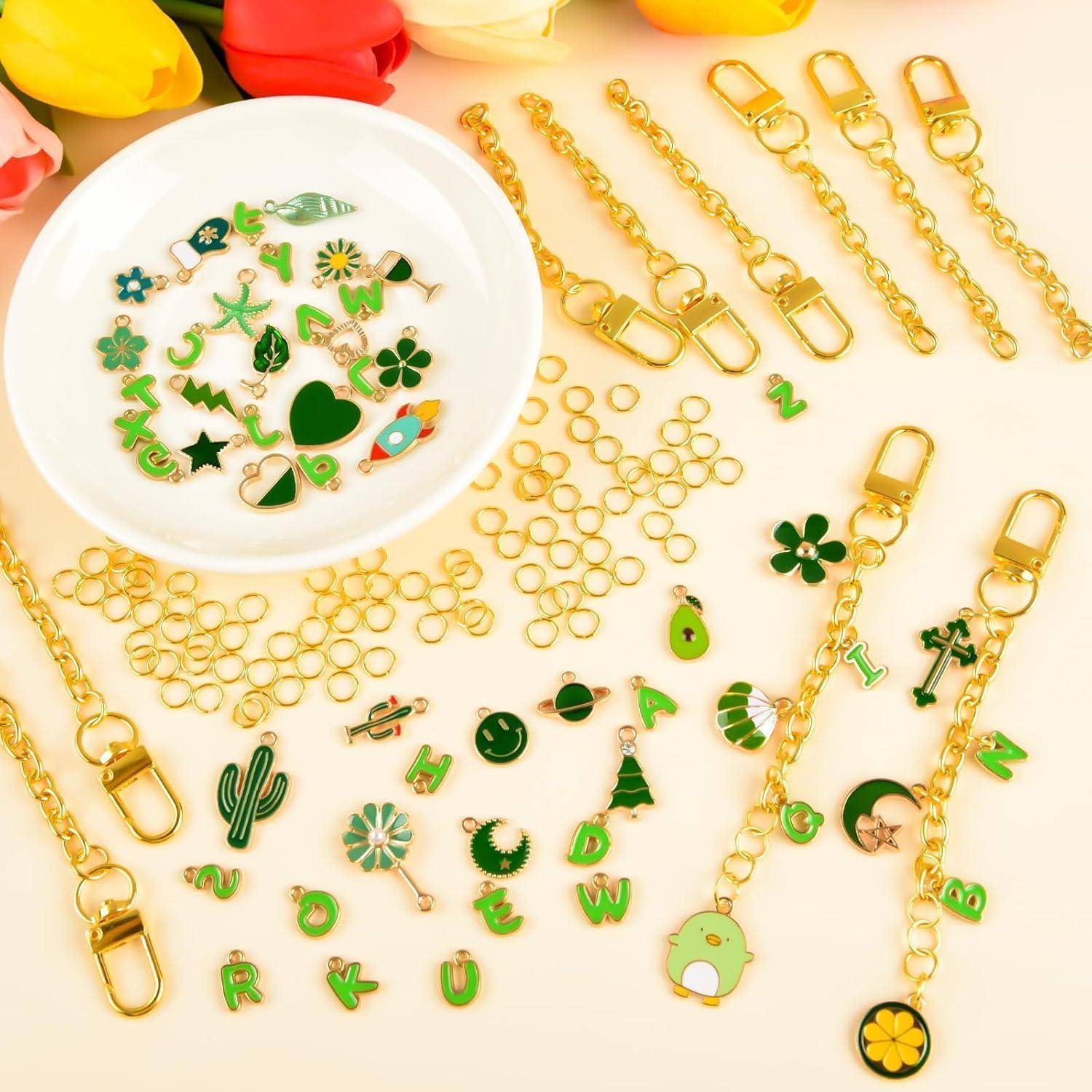 Sortierte goldene Schlüsselanhänger-Teile und grüne Buchstaben-Charms, die auf einer cremefarbenen Oberfläche verstreut sind, mit einer weißen Schale mit dekorativen Charms auf der linken Seite.