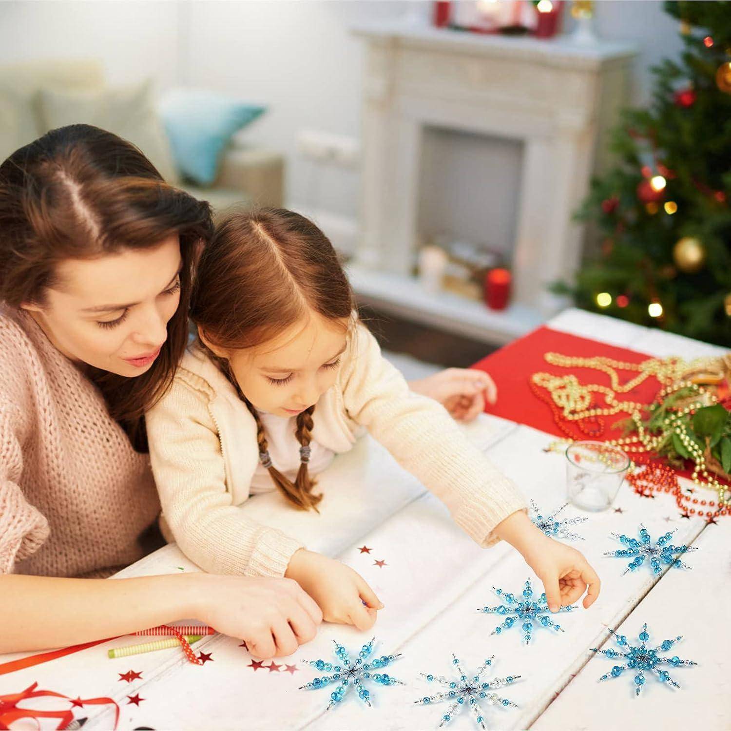 Mutter und kleine Tochter stellen Glitzer-Schneeflocken-Dekorationen an einem Tisch her, mit Weihnachtsdekoration und einem geschmückten Baum im Hintergrund.