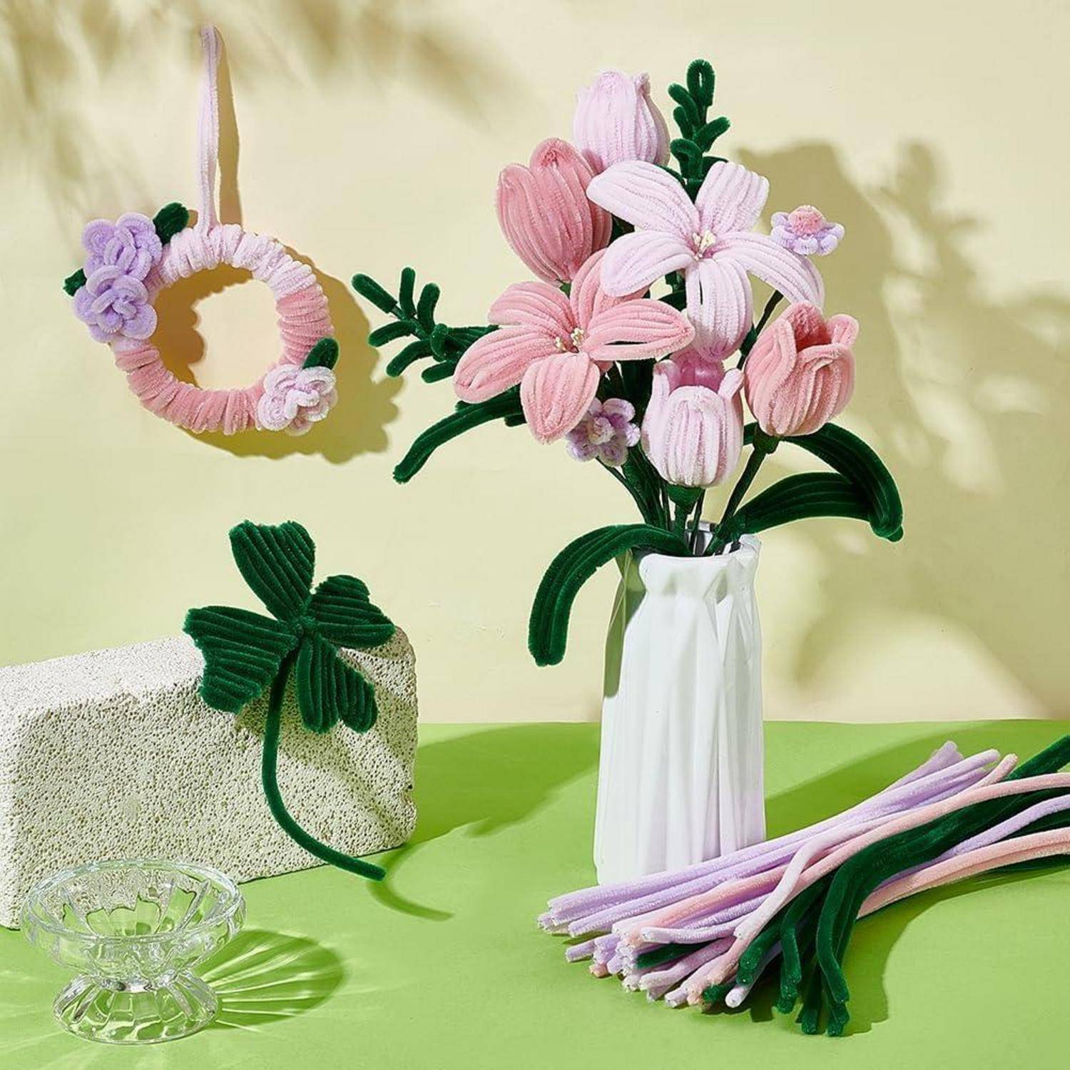 Pastellfarbene Garnblumen in einer weißen Vase mit passendem Garnkranz, Kleeblatt und Stielen auf einer grünen Tischplatte mit einer Glasschüssel angeordnet.