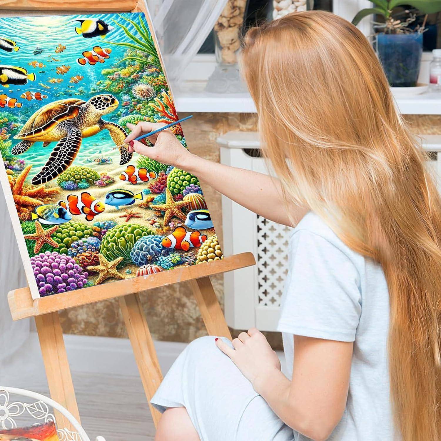Eine Frau malt eine farbenfrohe Unterwasserszene auf einer Leinwand, die eine Meeresschildkröte, einen Anemonenfisch, Korallen und einen Seestern auf einer Staffelei zeigt.