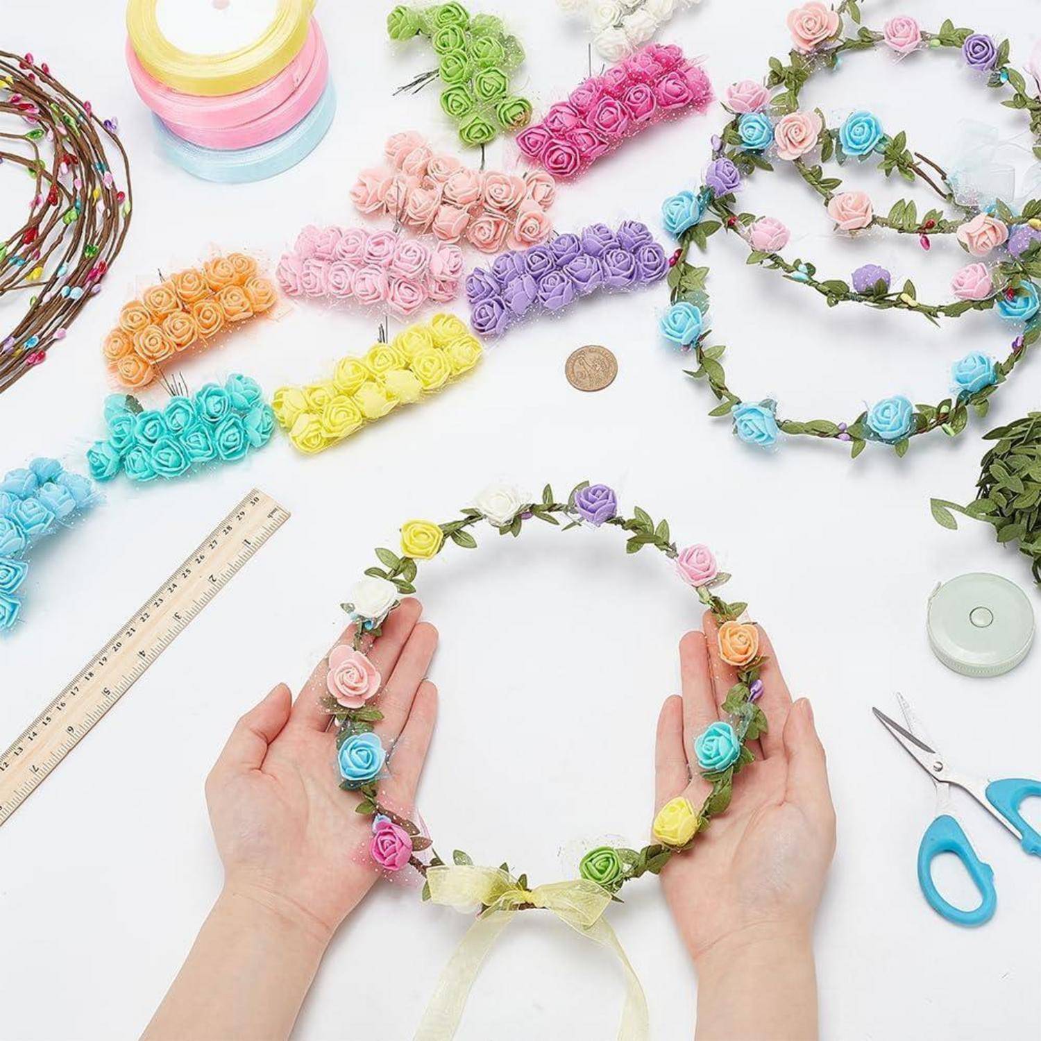 Hände halten ein Blumenstirnband aus zartem Rosenbutten mit Schleifenschleife; in der Nähe befinden sich farblich passende bunte Kränze, Bandrolle, Lineal, Schere und eine Münze auf einem Basteltisch.