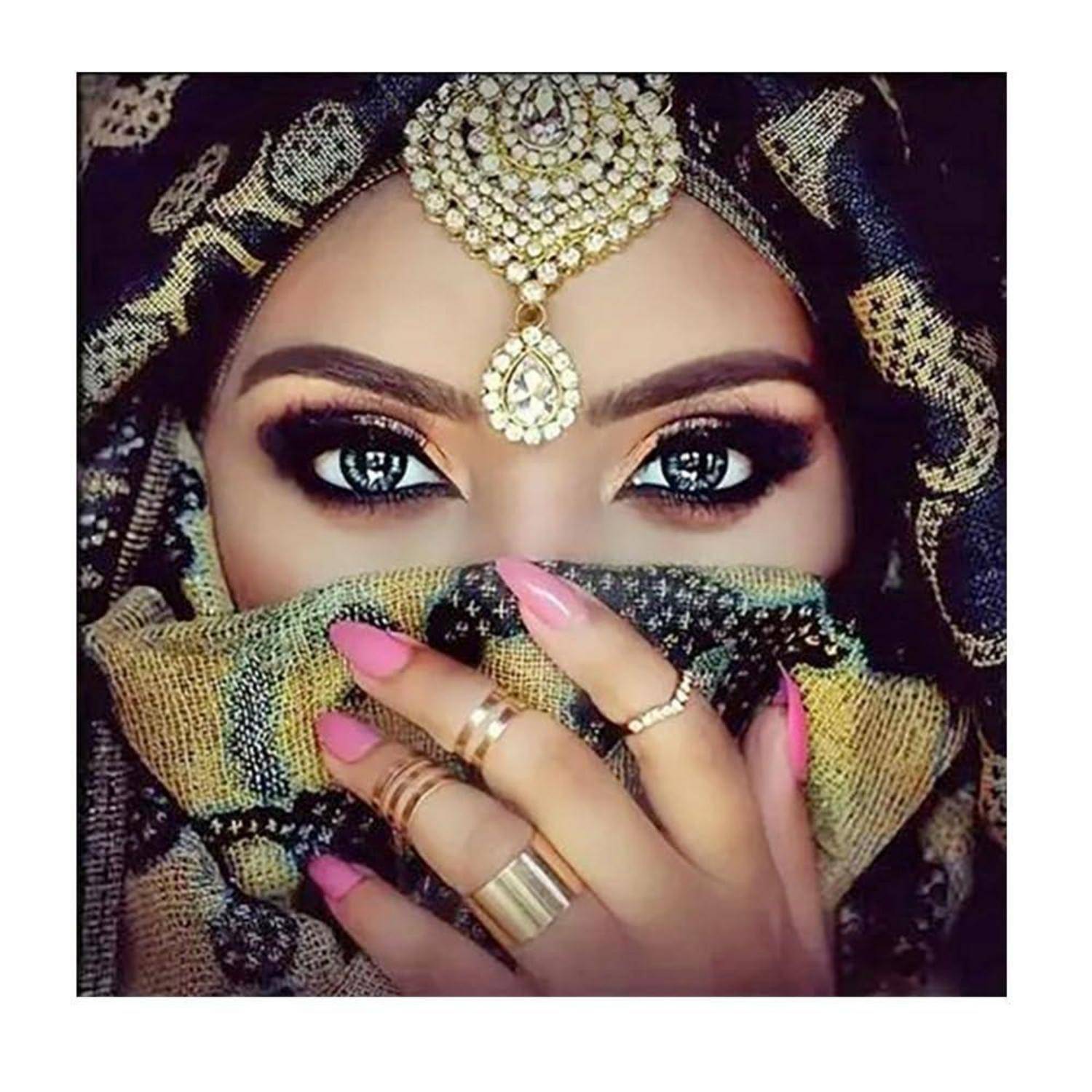 Nahaufnahme einer Frau mit aufwendigem goldenem Kopfschmuck und Schleier, der ihren Mund bedeckt, zeigt dramatisches Augen-Make-up und rosa manikürtere Hand mit Ringen.