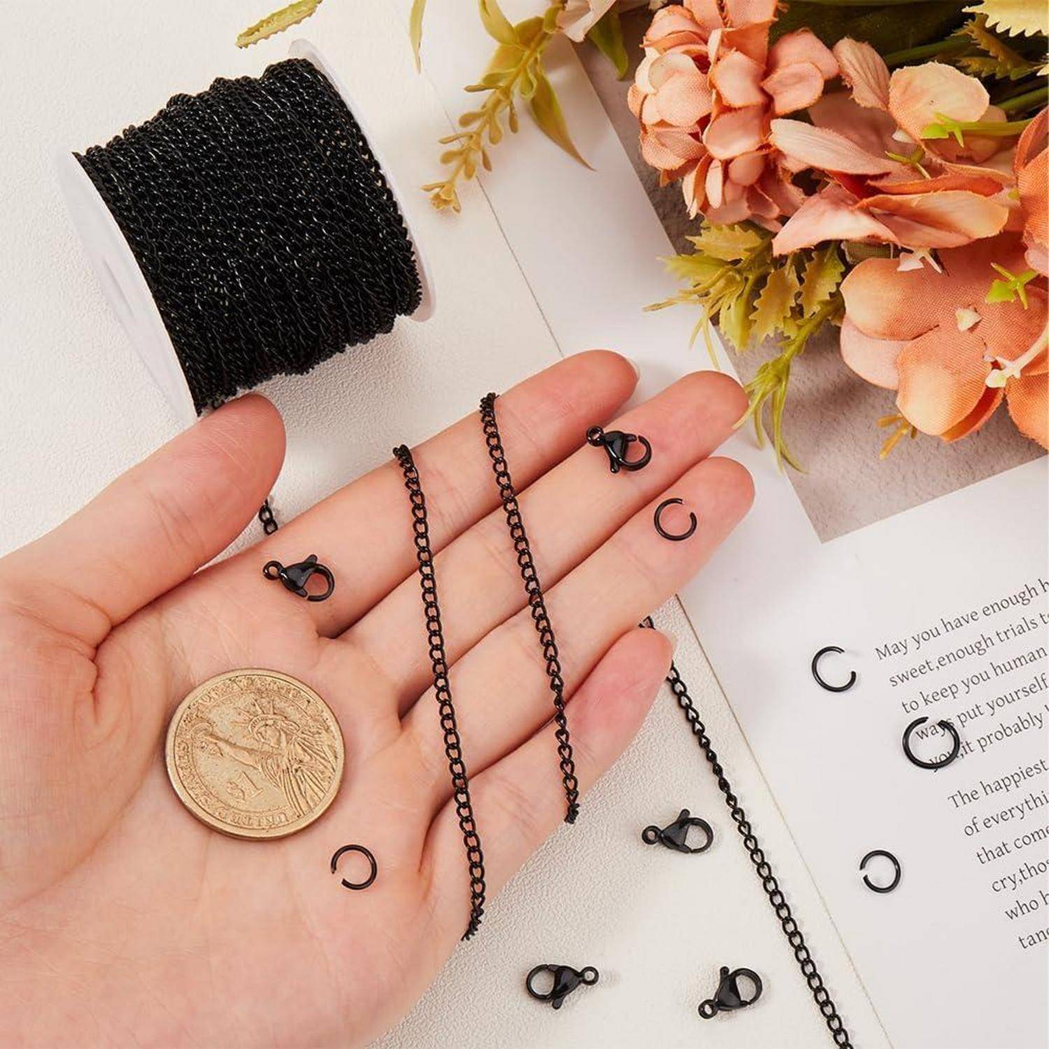 Hand hält schwarze Kette und kleine Schmuckspangen neben einer Spule Schnur, Münze und Blumen auf einer Handwerksfläche.