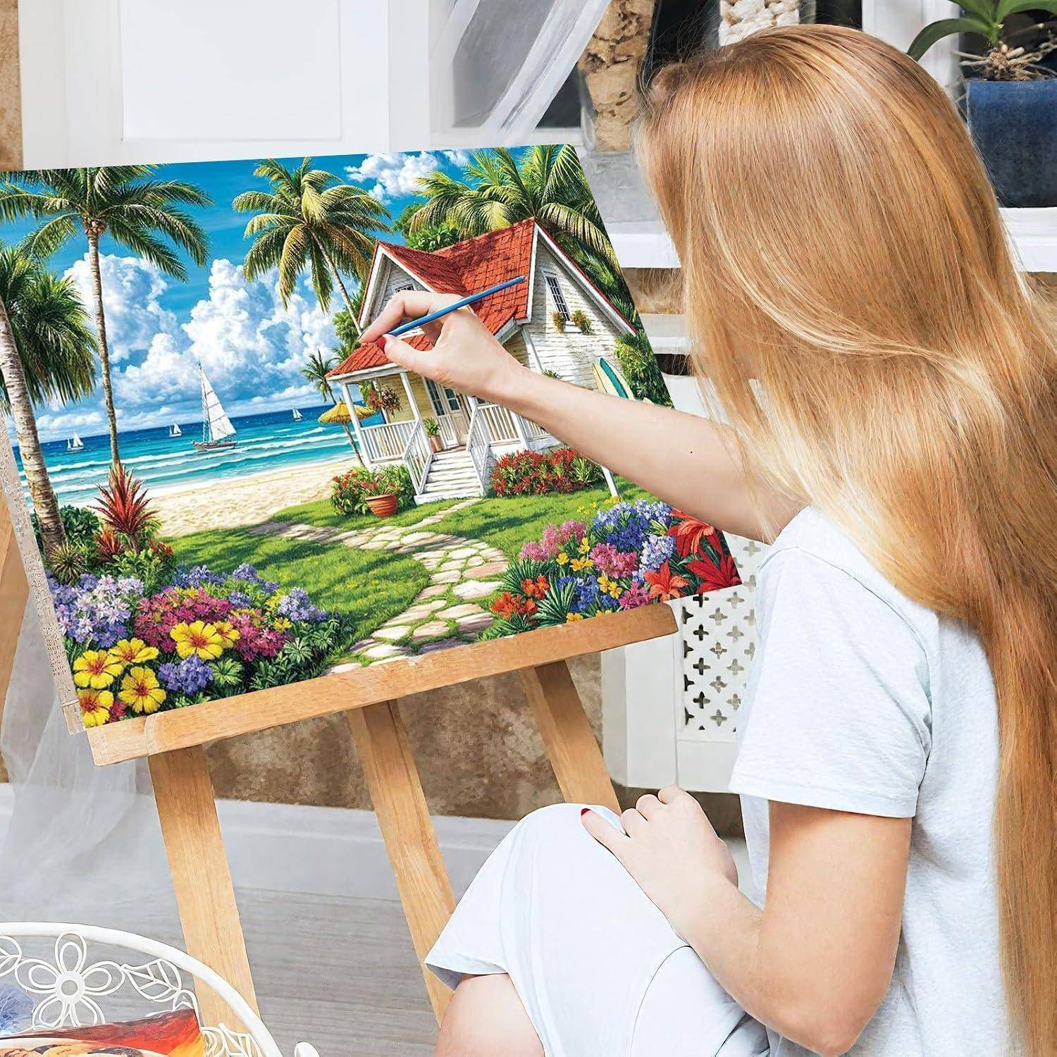 Eine Frau malt eine bunte tropische Strandszene auf einer Staffelei, mit Palmen, Blumen, einem Haus und Segelbooten am Meer.