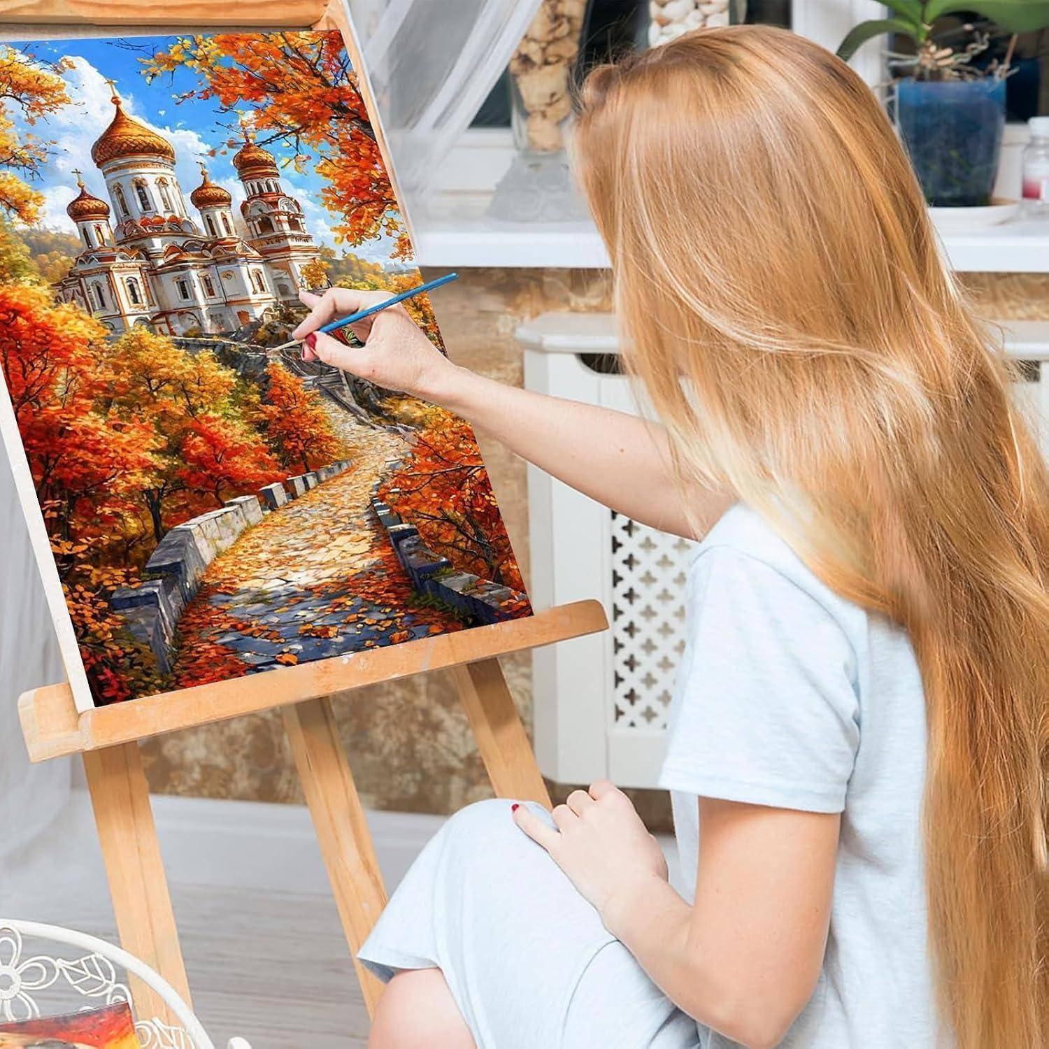 Eine Frau malt eine detaillierte Herbstlandschaft mit einer Kathedrale auf einer Staffelei in Innenräumen.