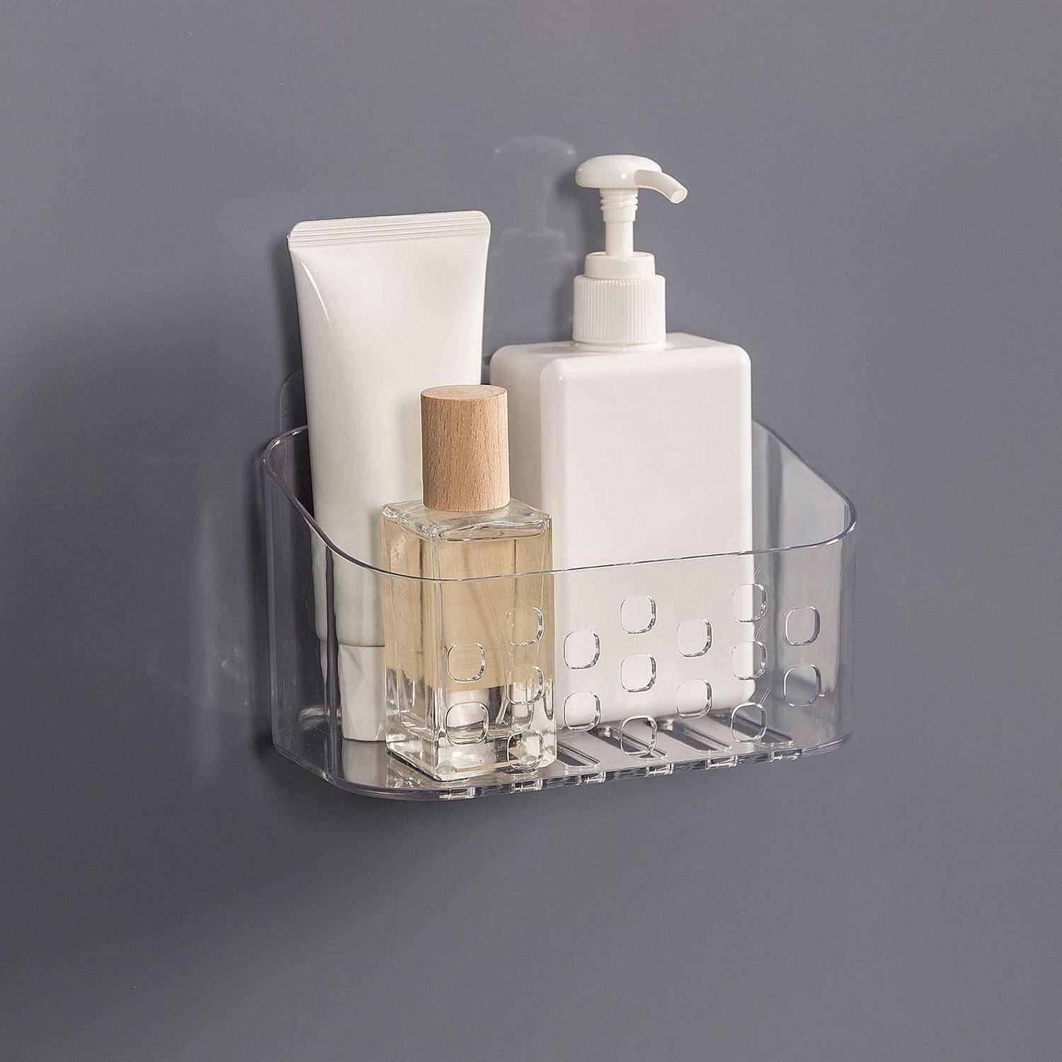 Transparenter, an der Wand befestigter Badorganizer mit einer weißen Lotionspumpe, einer Tube und einer kleinen Glasflasche mit Holzverschluss an einer grauen Wand.