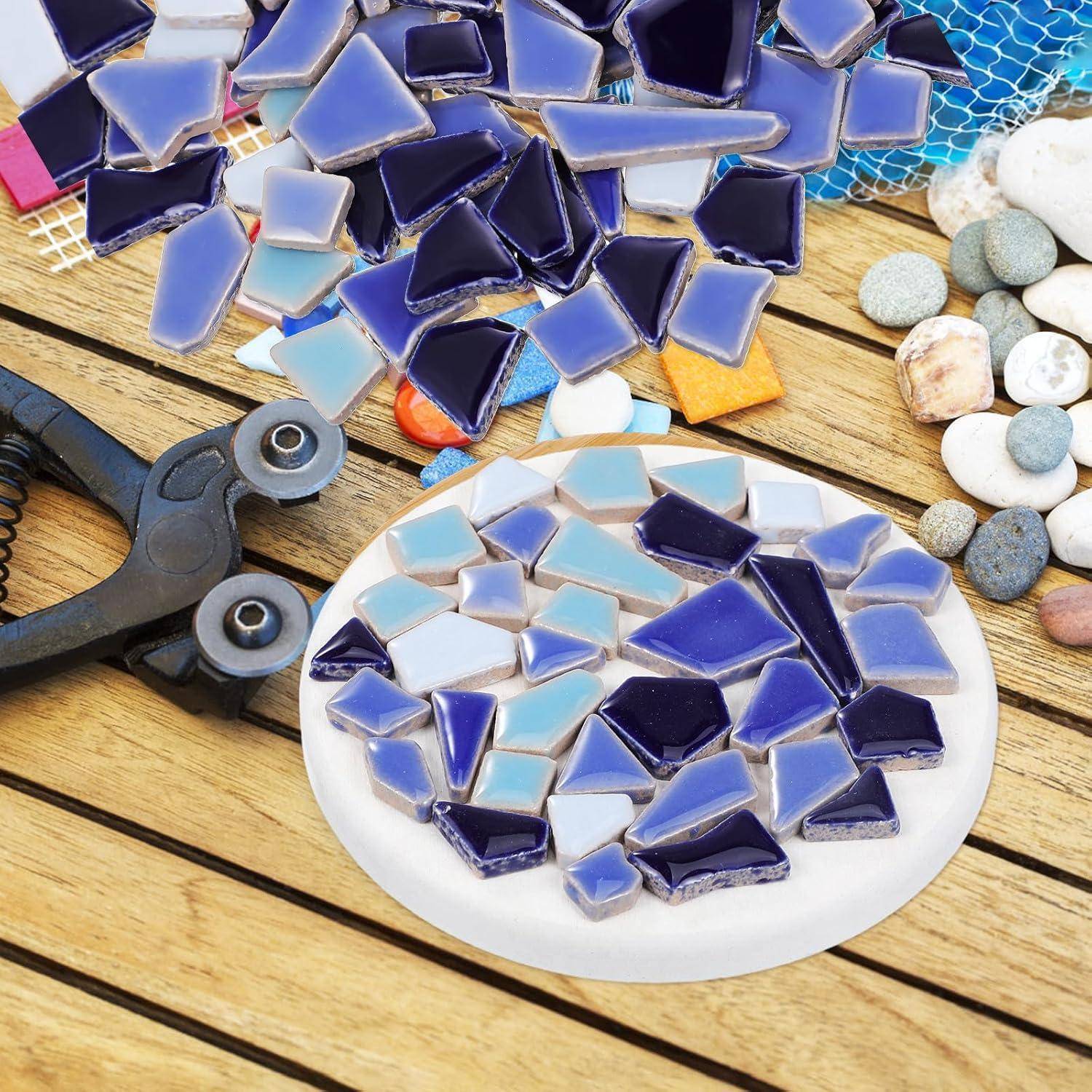 Sortierte blau-weiße Mosaikglaskacheln auf einer weißen runden Basis, mit Fliesenknabber und Dekorsteinen auf einer Holzarbeitsfläche.