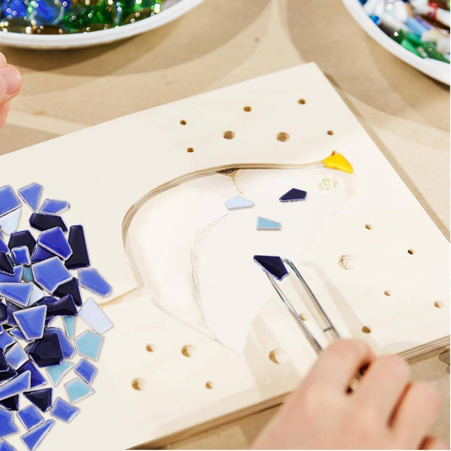 Hände arrangieren blaue Mosaikfliesen auf einer weißen Tafel und bilden eine gebogene Form auf einem Handwerkstisch.