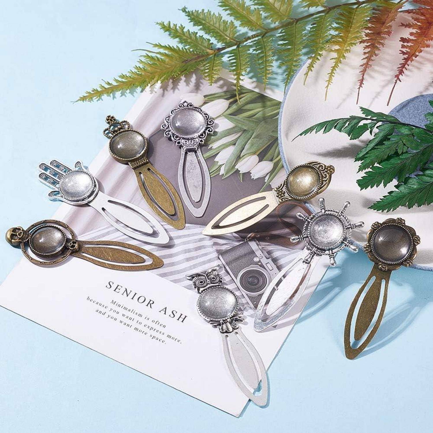 Dekorative Metall-Haarspangen in Tropfenform in Silber und Gold, angeordnet auf einer Magazinseite mit grünen Farnblättern.