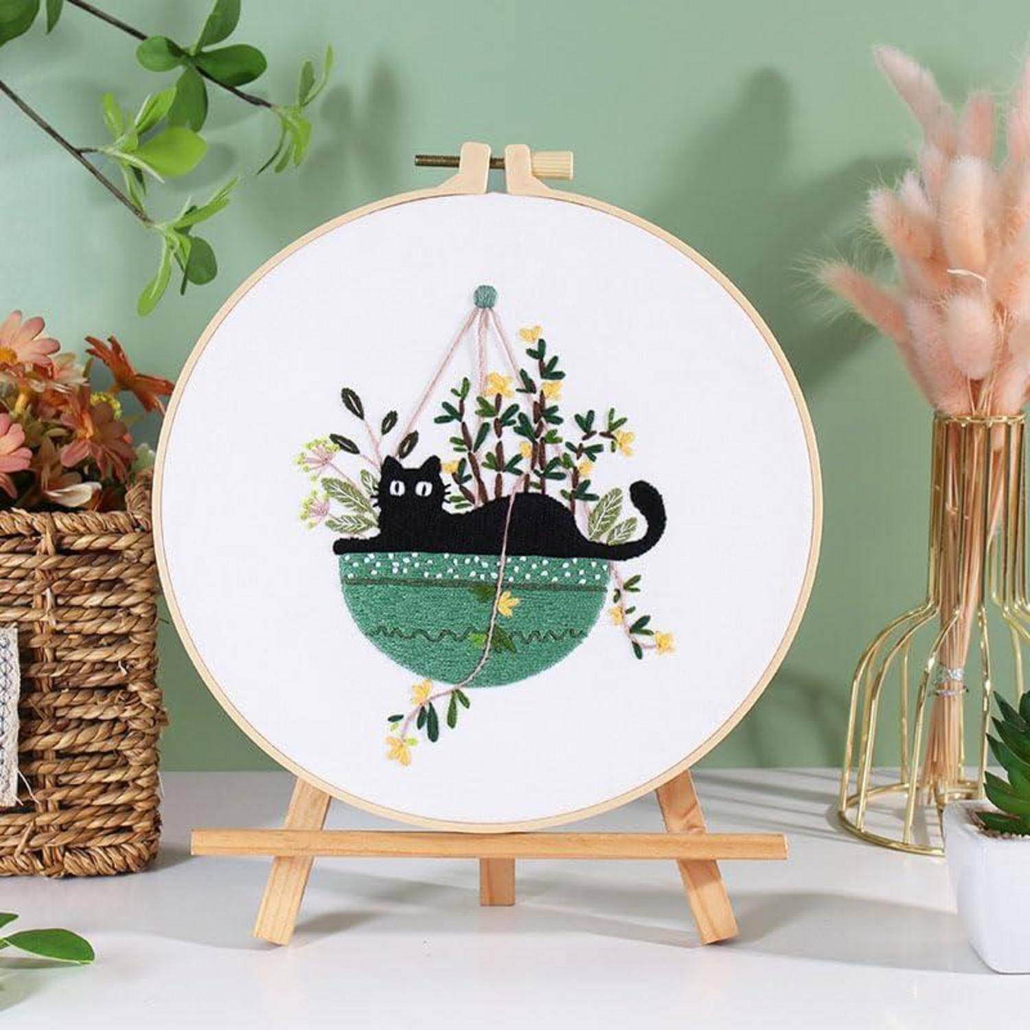 Ein Stickrahmen auf einer kleinen Holzstaffelei zeigt eine schwarze Katze, die in einem grünen hängenden Blumenkasten mit Blumen und Blättern liegt.