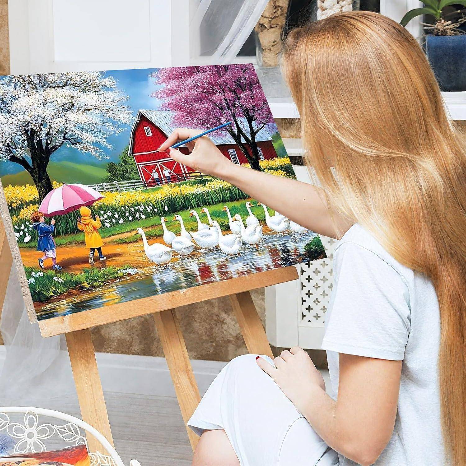 Eine Frau malt eine bunte Landszene auf eine Leinwand, mit einer roten Scheune, Bäumen, Kindern unter einem Regenschirm und weißen Gänsen in der Nähe von Wasser.