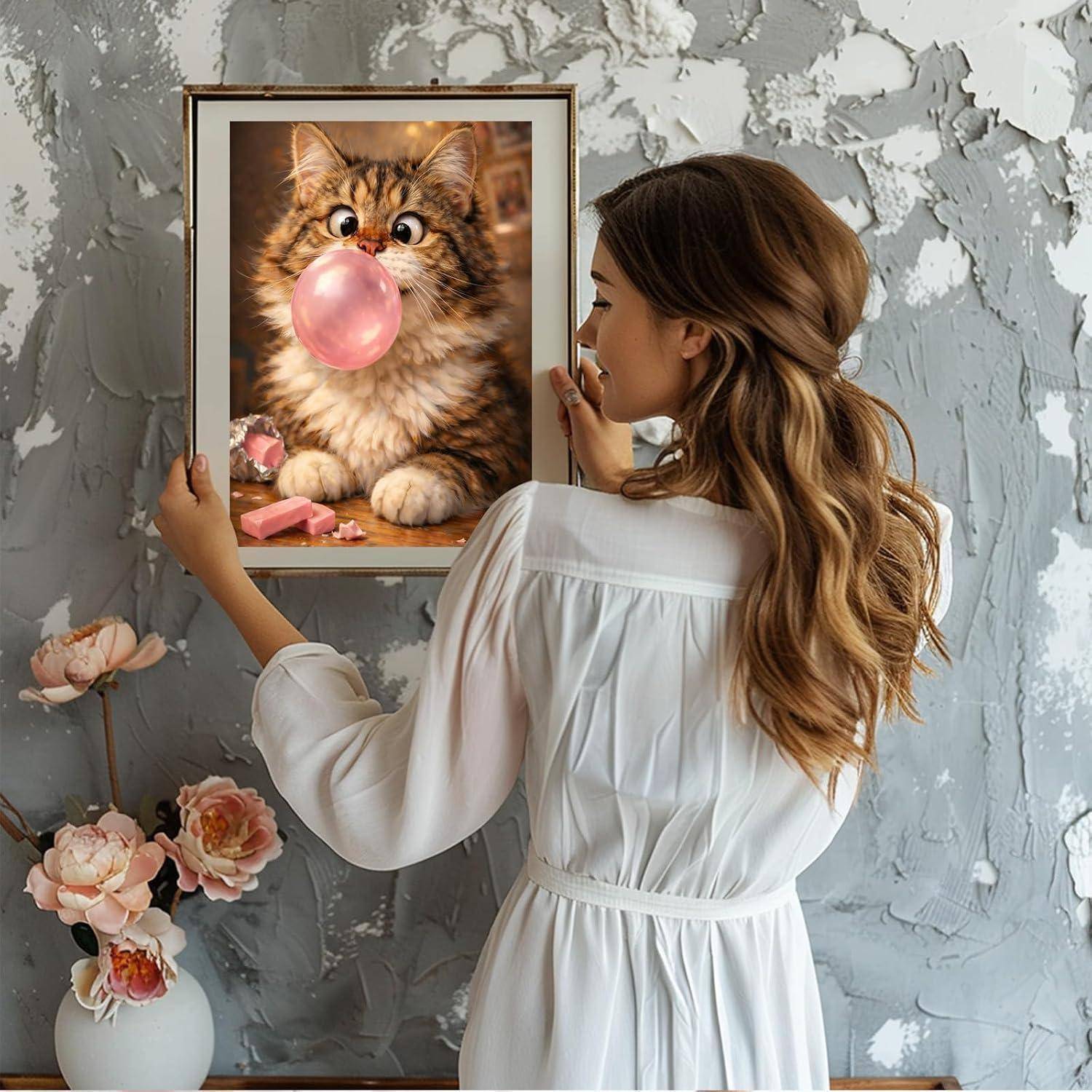 Eine Frau in weißem Kleid hängt ein gerahmtes Bild einer flauschigen Katze auf, die rosa Kaugummi bläst, an einer strukturierten grauen Wand auf, mit Blumen darunter.