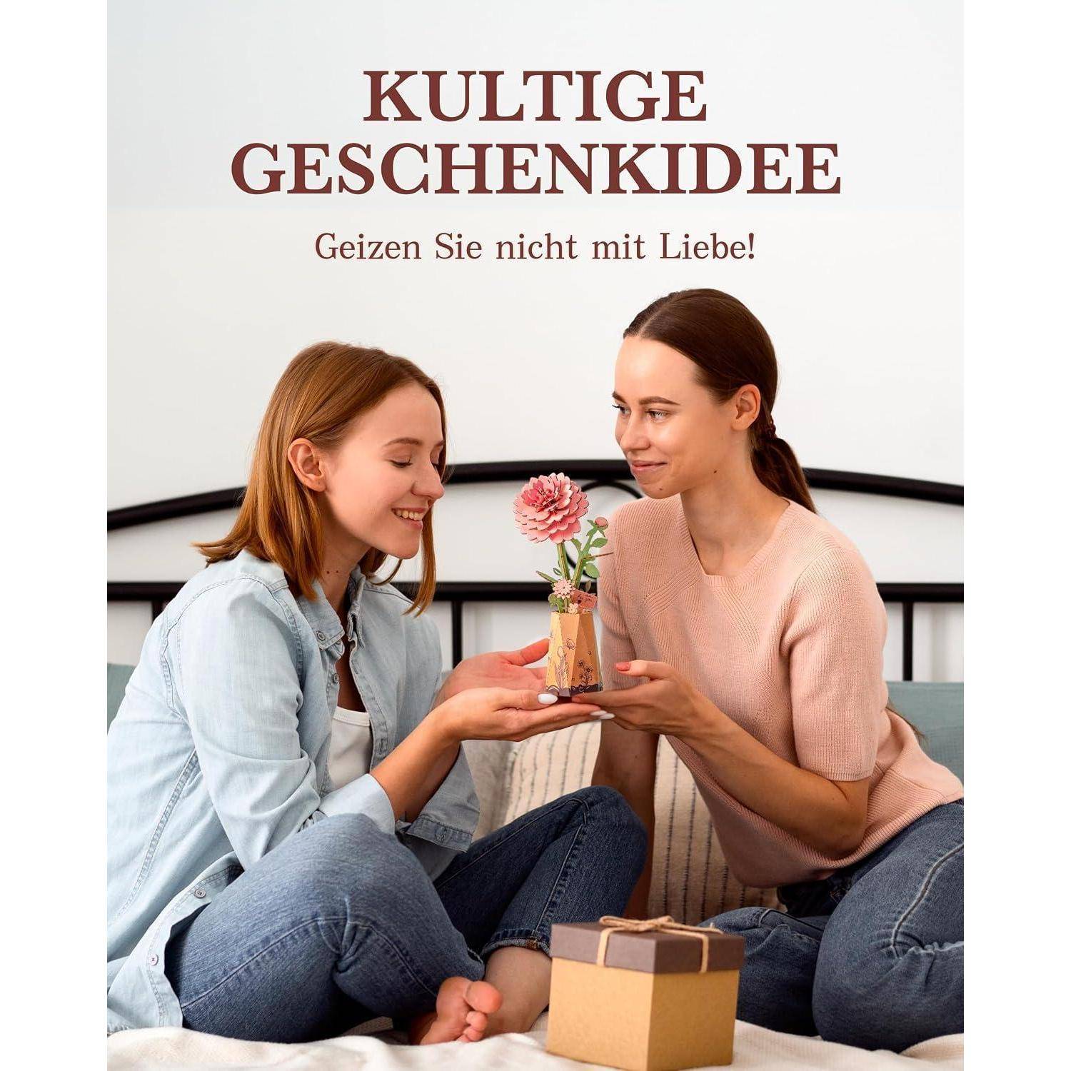 Zwei Frauen sitzen auf einem Bett und tauschen ein kleines Geschenk und eine rosa Blume aus; der Text lautet „KULTIGE GESCHENKIDEE