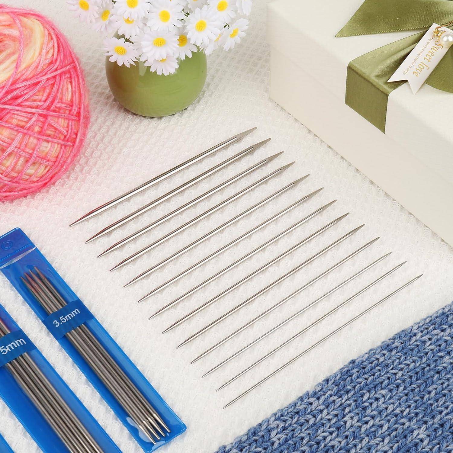Satz von Metallstricknadeln nach Größe auf einer weißen Oberfläche angeordnet, mit blauen Nadelpackungen, Garn, Blumen und einer Geschenkbox in der Nähe.