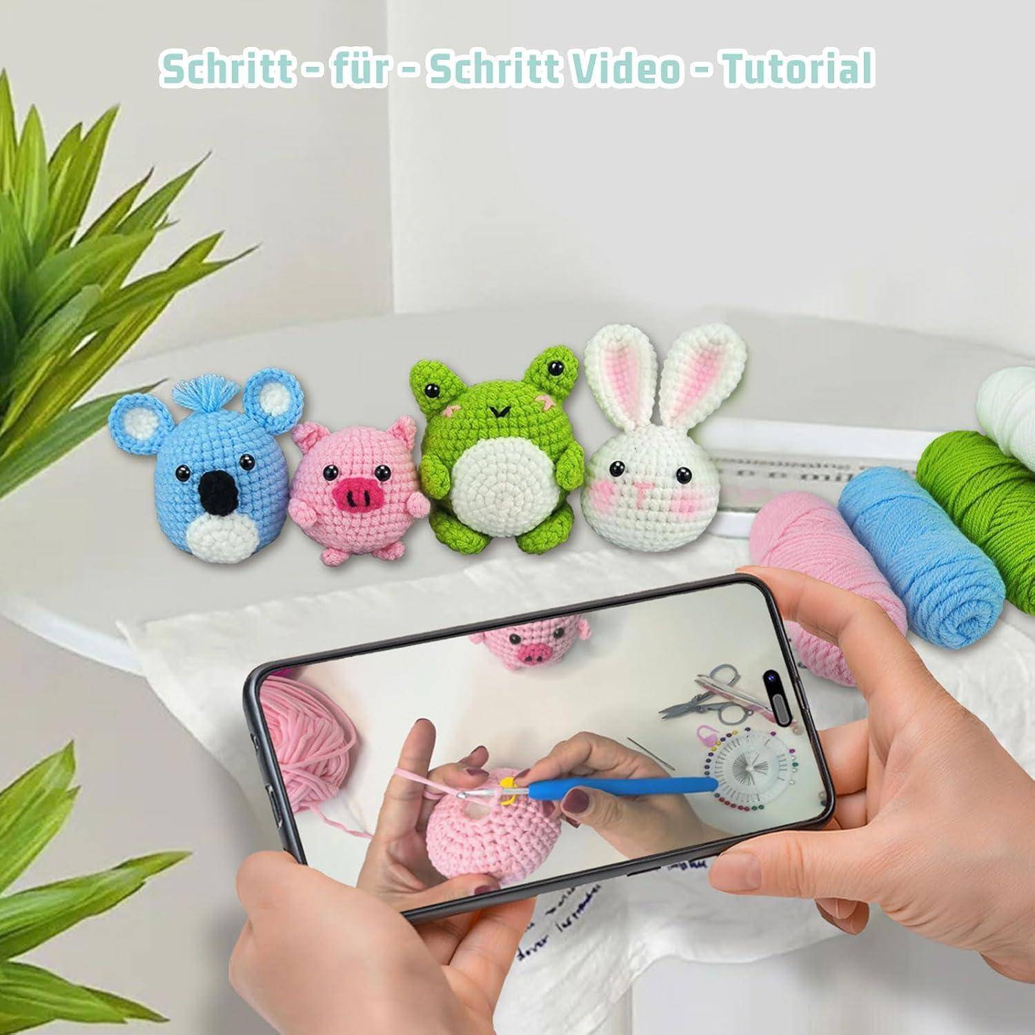Hände, die ein Smartphone verwenden, um ein Häkel-Tutorial aufzunehmen, mit fertigen Amigurumi-Tieren und Garn auf einem weißen Tisch.