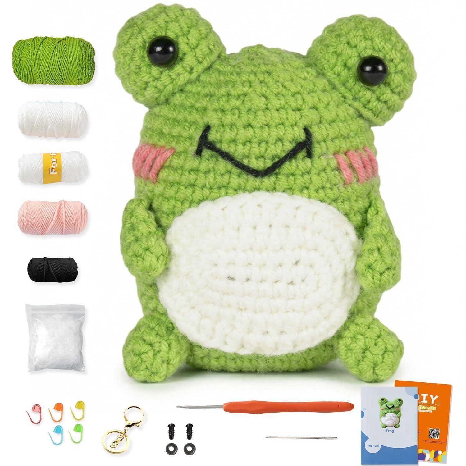 Gehäkelte grüne Froschplüsch mit weißem Bauch und schwarzen Perlenaugen, gezeigt mit Garn, Häkelhaken, Sicherheitsaugen und Kitverpackung.
