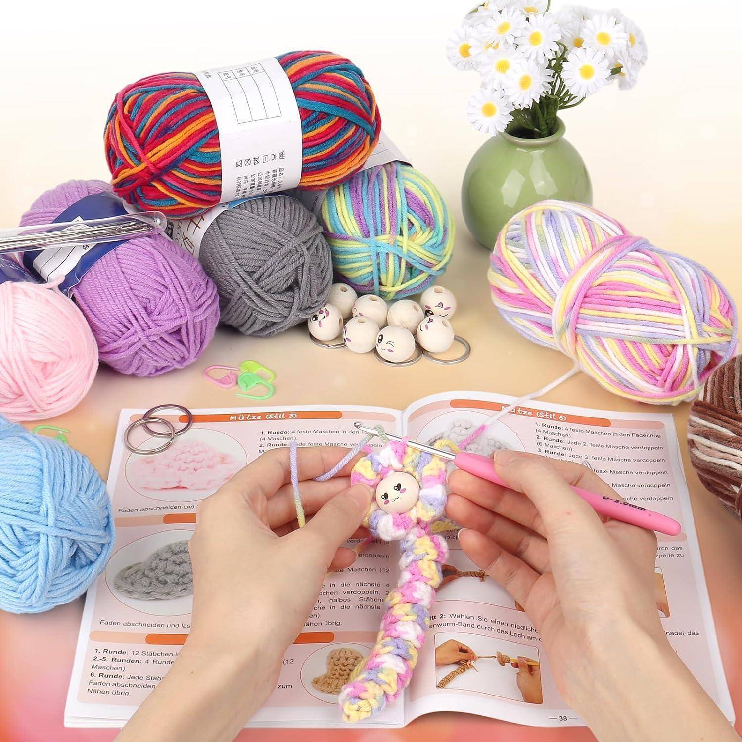 Hände häkeln einen bunten Amigurumi-Anhänger auf einem offenen Musterbuch, umgeben von Wollknäueln, Häkelnadeln, Maschenmarkierern und einer Vase mit Blumen.